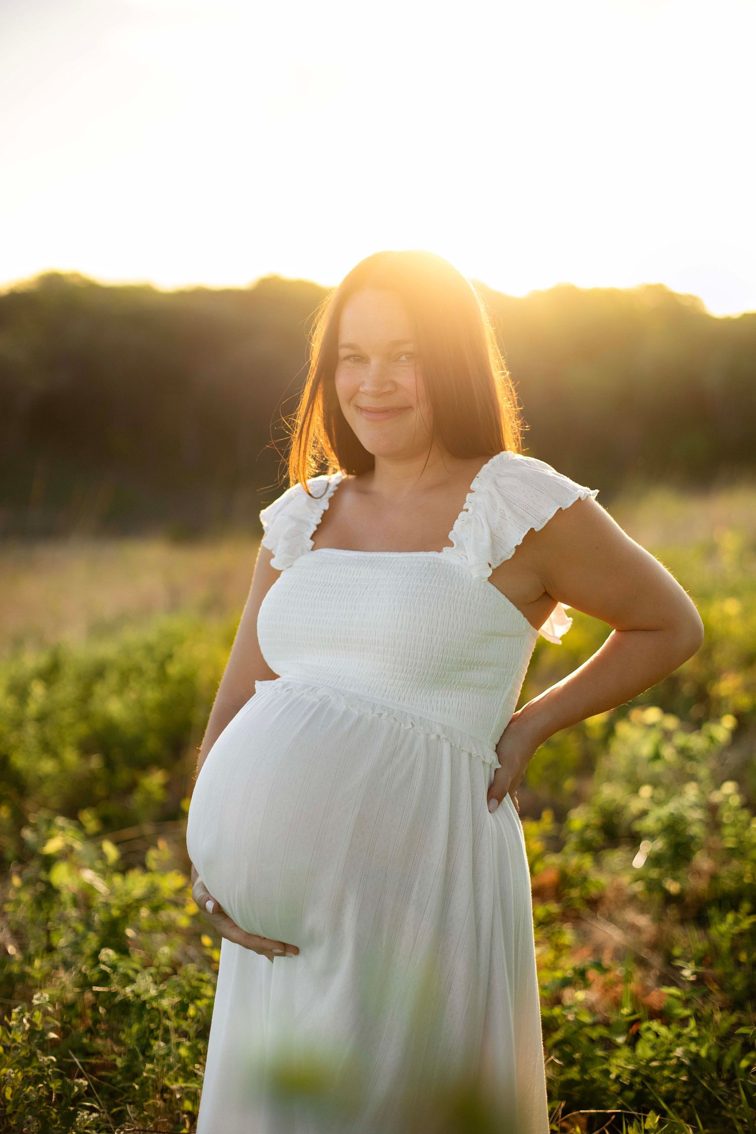 Pregnant woman in white dress smiling while holding baby bump in sunny field.