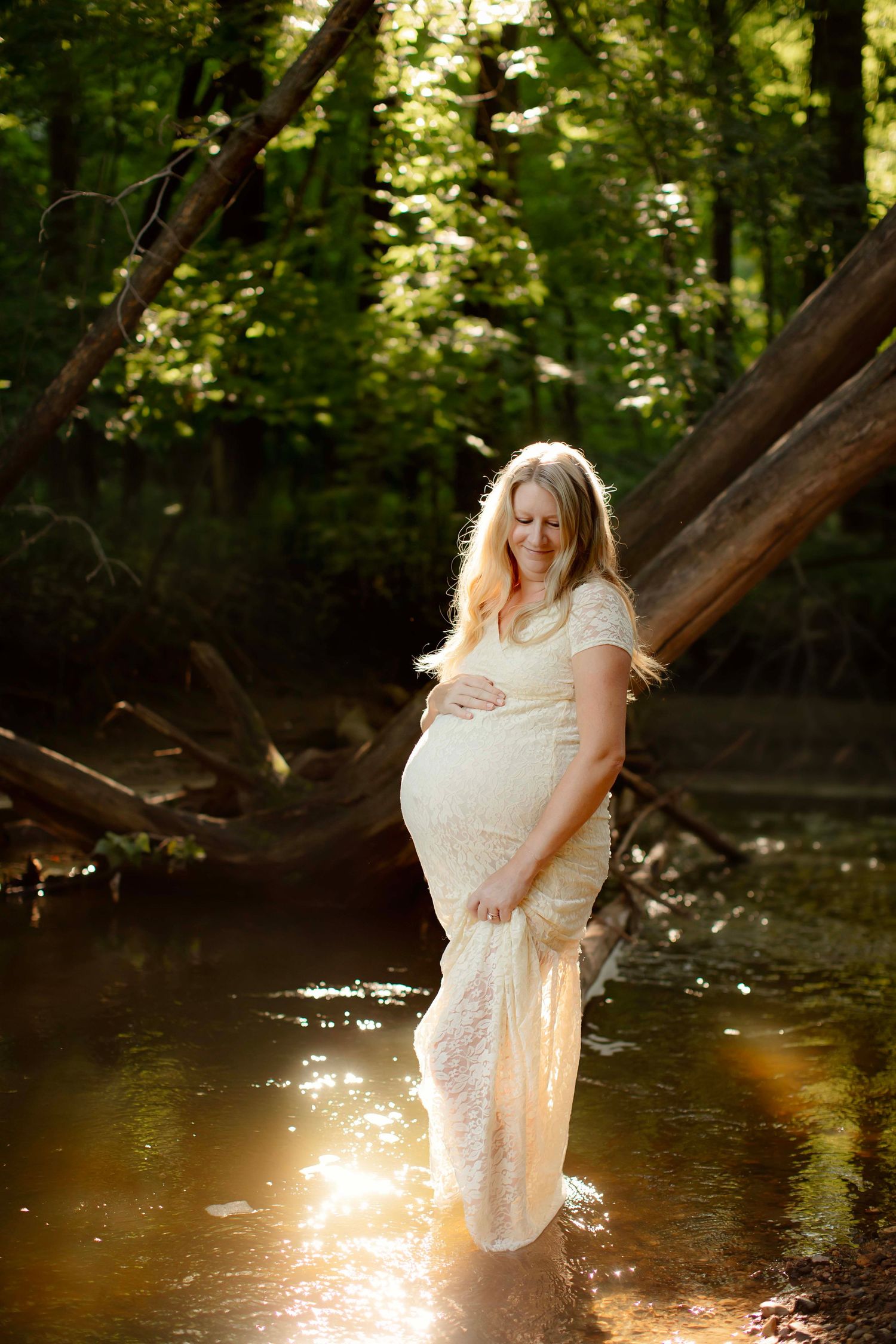 Pregnant woman in white lace dress standing in sunlit forest creek, cradling baby bump surrounded by lush green trees.