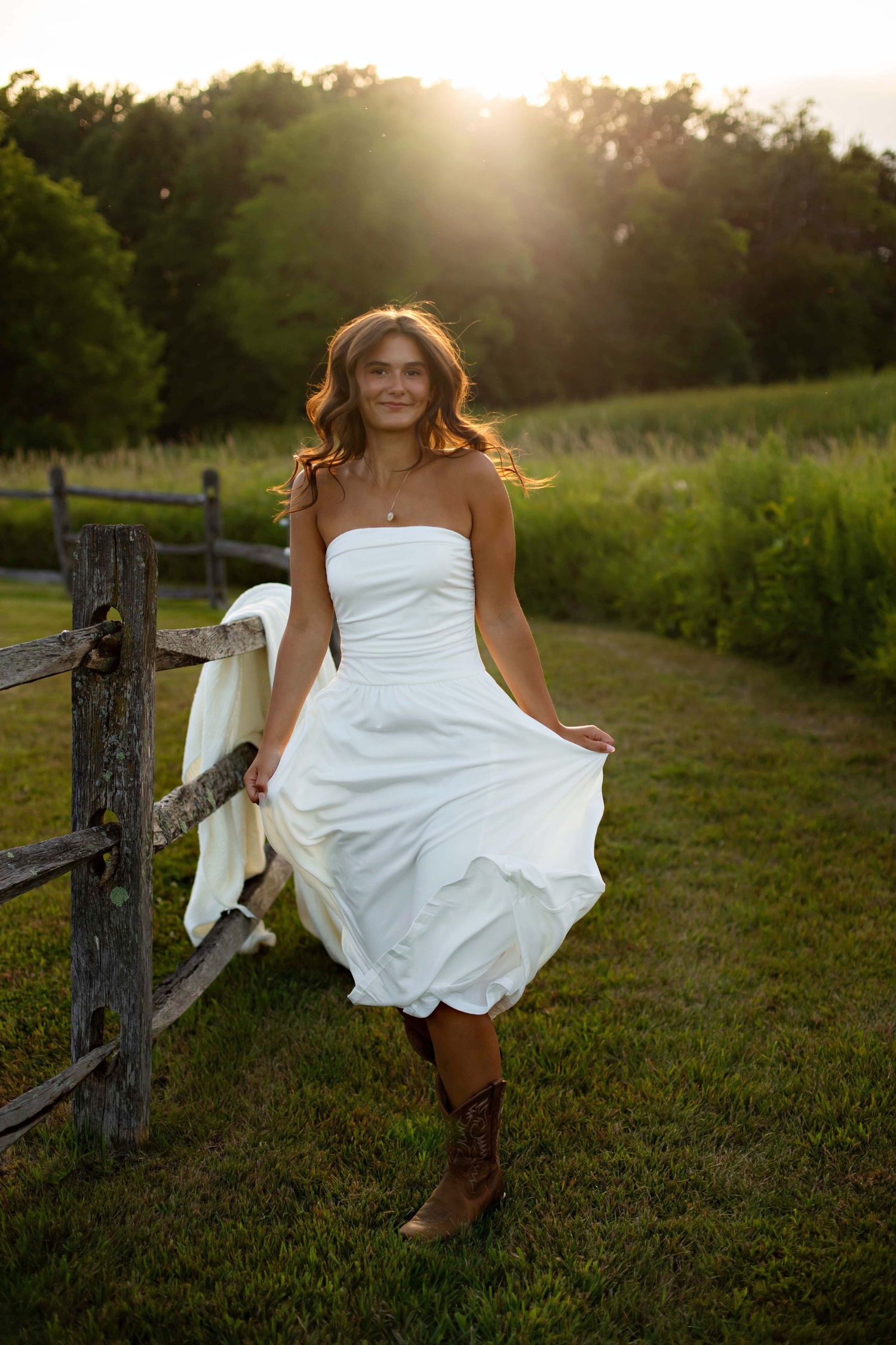 Woman in white strapless dress and cowboy boots leaning on rustic fence at golden hour sunset.