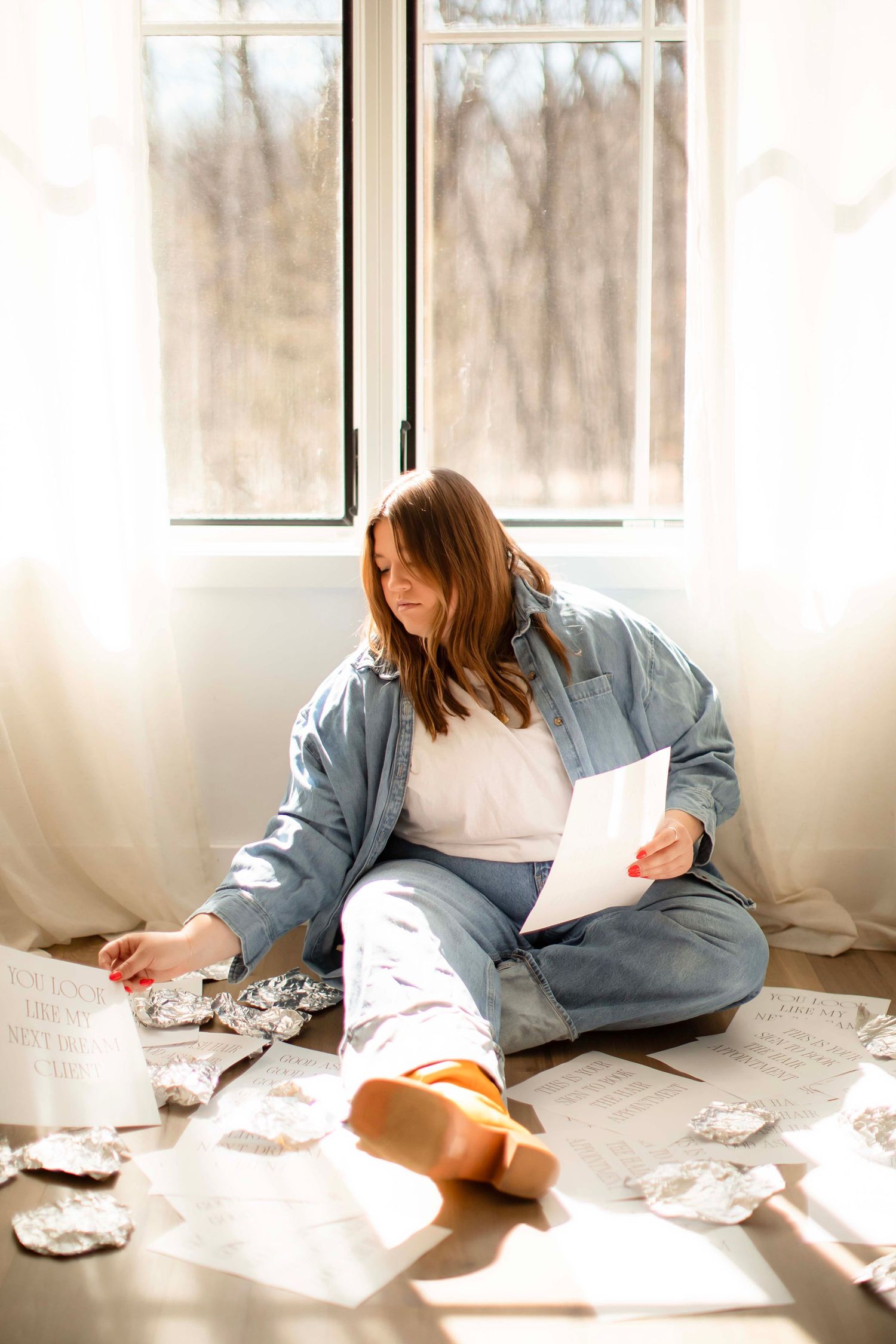 Hair stylist  in denim jacket sits on floor by sunny window, reading papers surrounded by hair foils