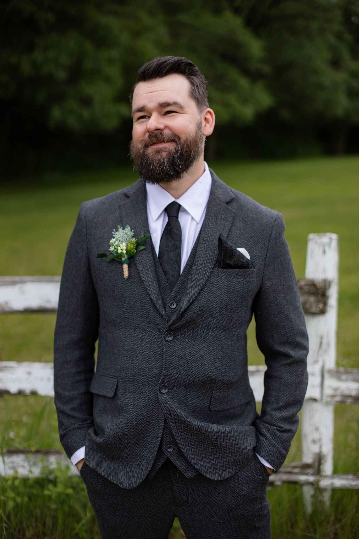 A person in a dark gray suit and tie smiles while standing outdoors against a blurred natural background.