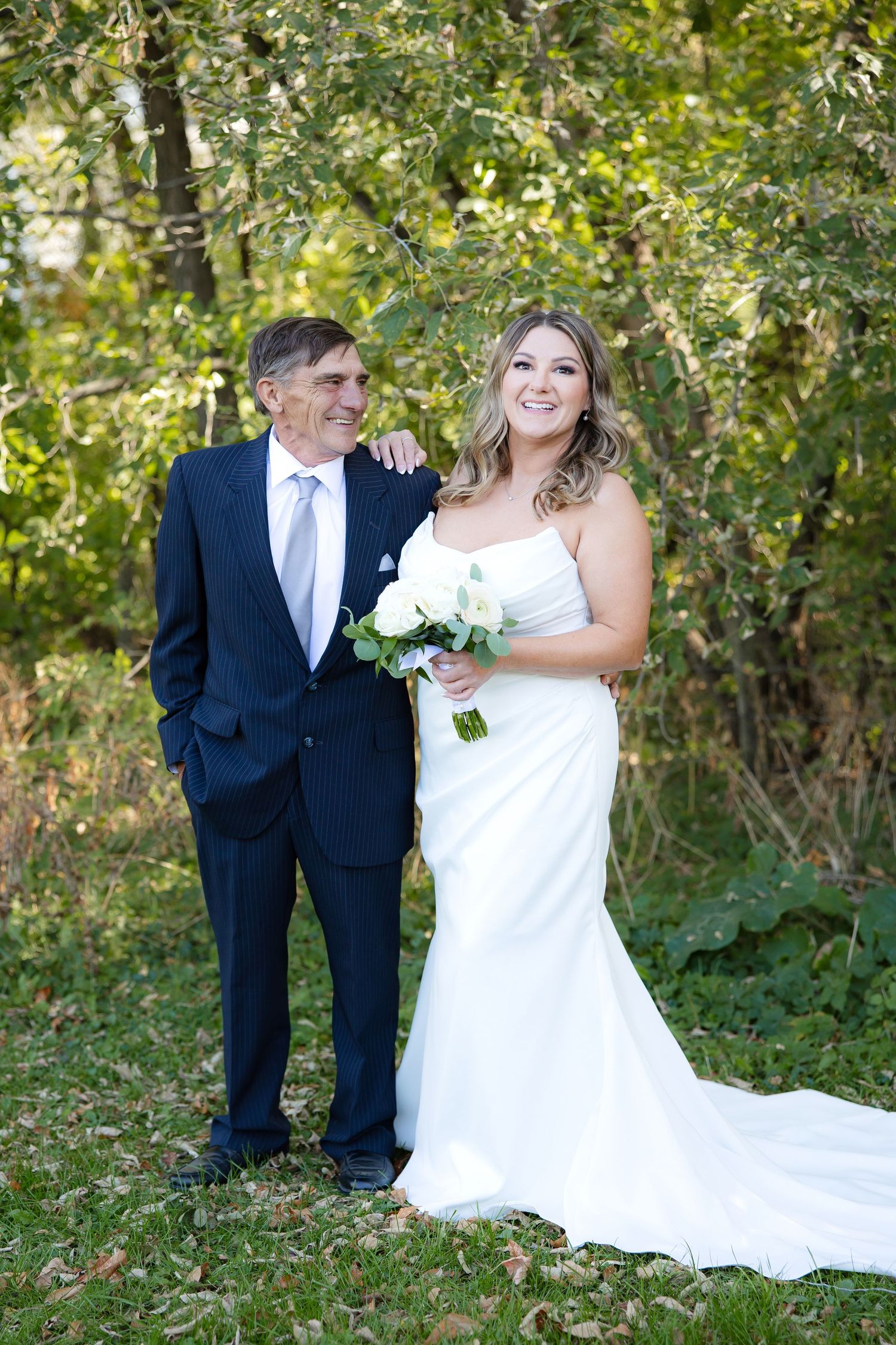Bride in flowing white dress poses for outdoor wedding portraits with attendants in garden setting.