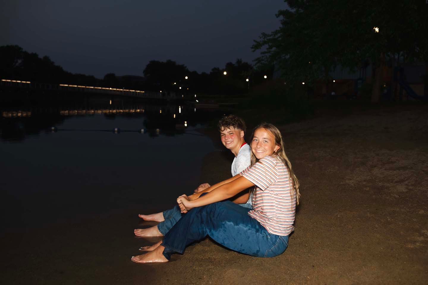 Two young people sitting together on a sandy shore by a calm lake at night with lights reflecting on water.