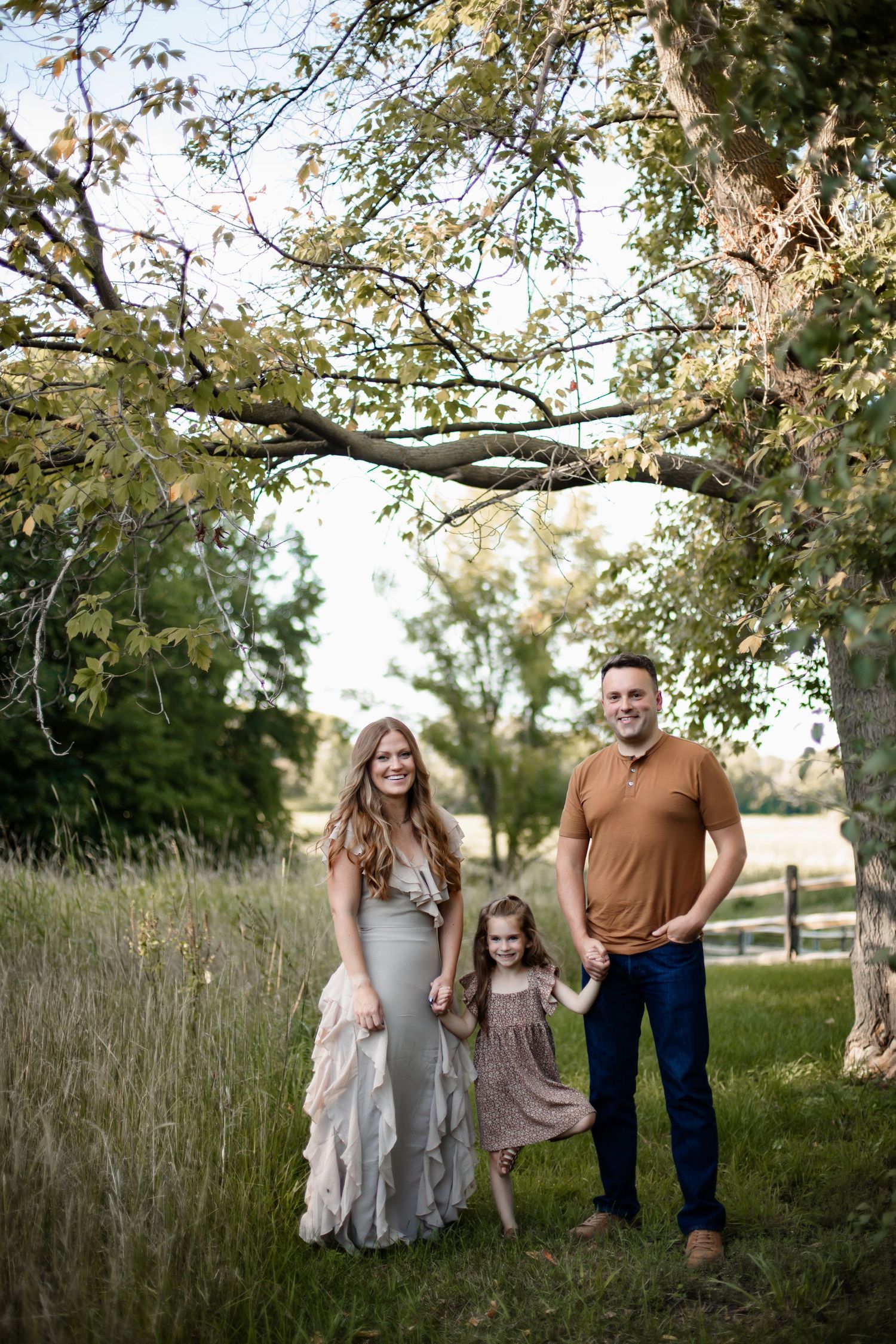 A family stands together in a scenic outdoor setting with tall grass and trees on a sunny day.
