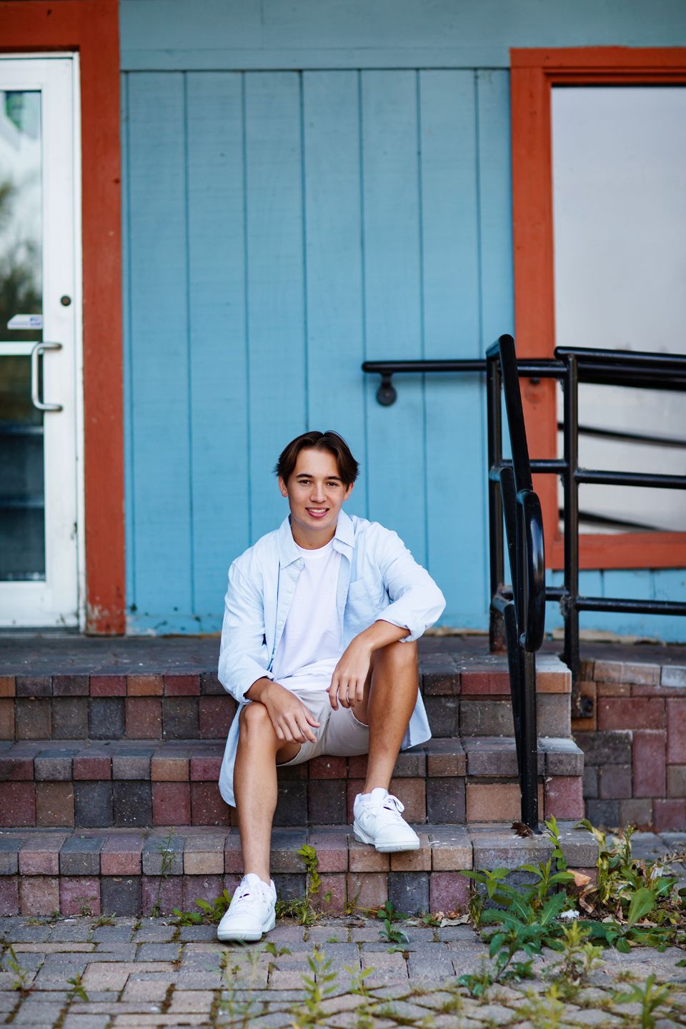 Casual portrait of someone seated on brick steps wearing white sneakers and a light-colored outfit against a blue wall.