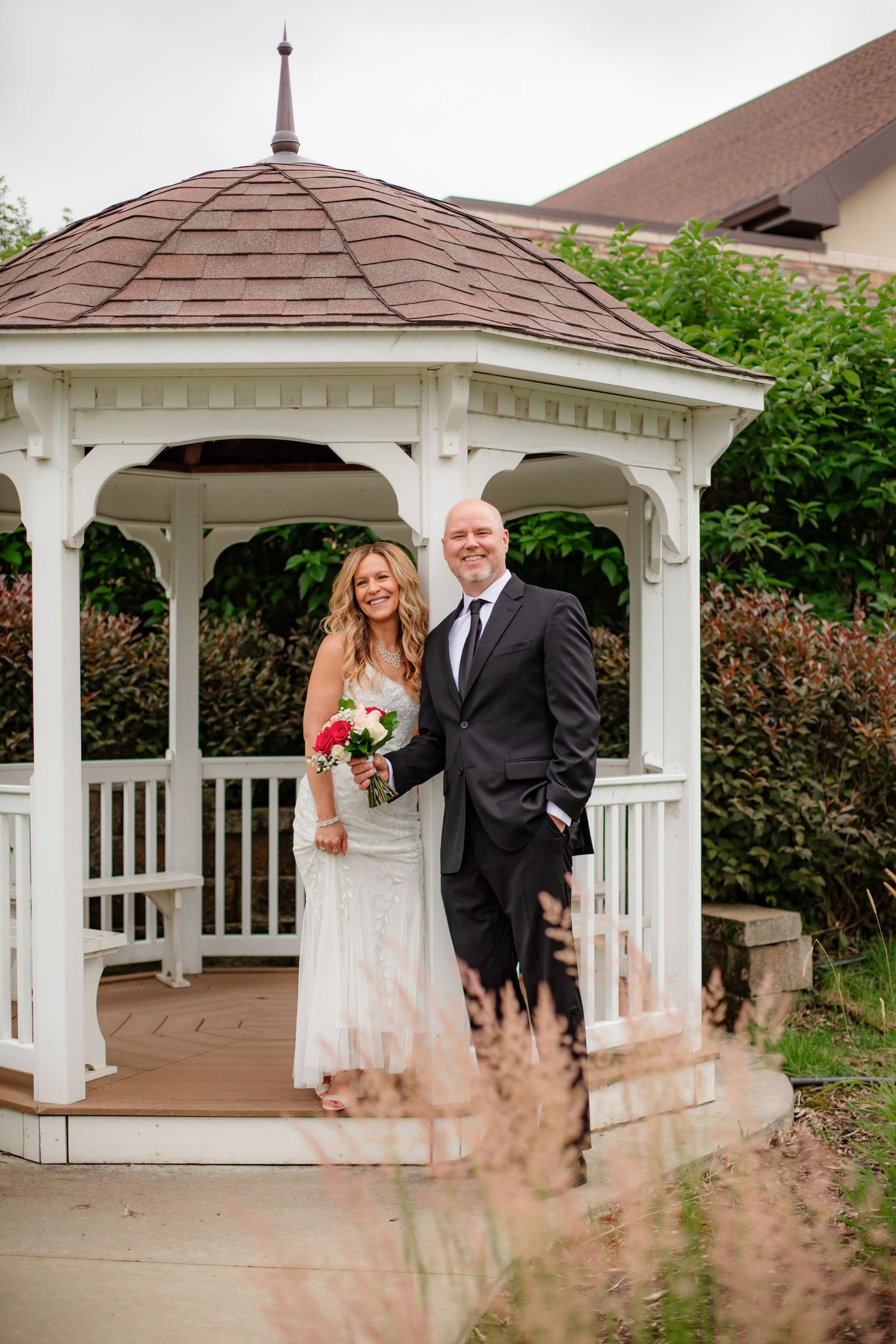 White wooden gazebo with decorative trim and brown shingled roof in a garden setting.