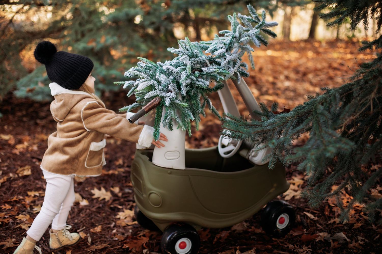 Child placing holiday decorations on an olive green toy car in a forest setting on autumn day.