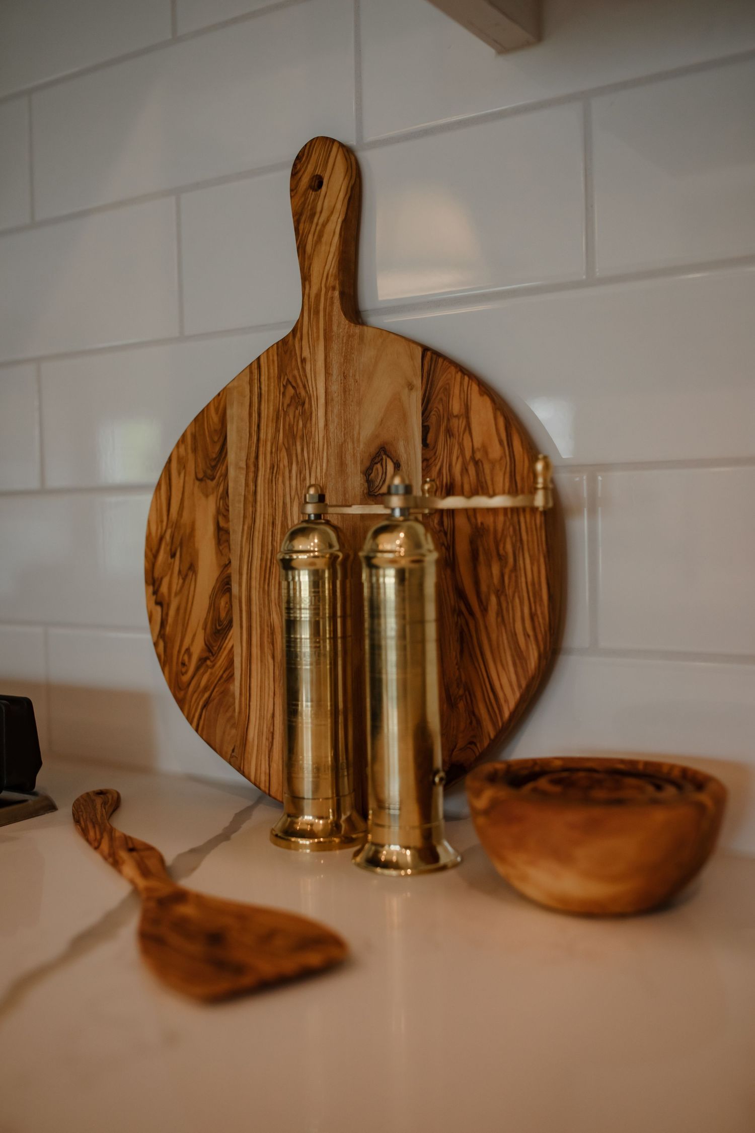 Wooden cutting board with brass pepper mills and wooden bowls against white tile backsplash.