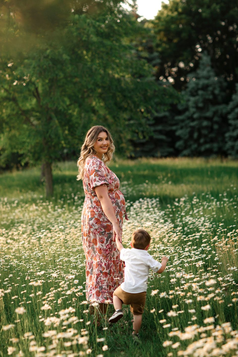 In a golden meadow of daisies, a woman in a pink floral dress walks with a small child among blooming flowers.