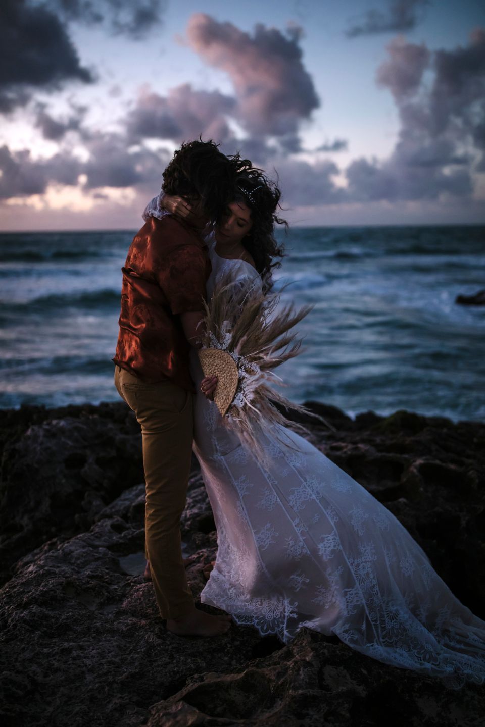 A couple shares an intimate moment in wedding attire against a moody oceanside backdrop at dusk.