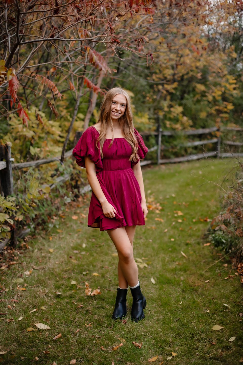 Burgundy dress and black boots create striking contrast against fall foliage and rustic fence background.