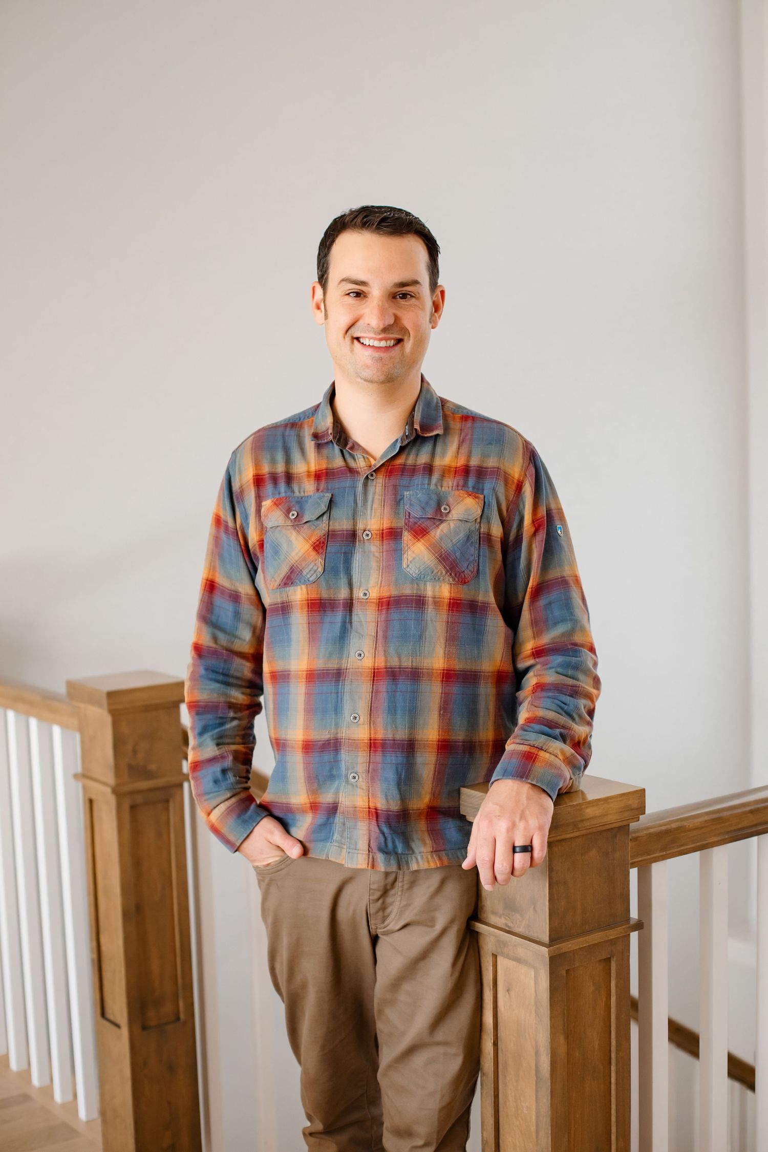 Professional portrait of someone in a colorful plaid button-up shirt and khaki pants standing by wooden stairs.