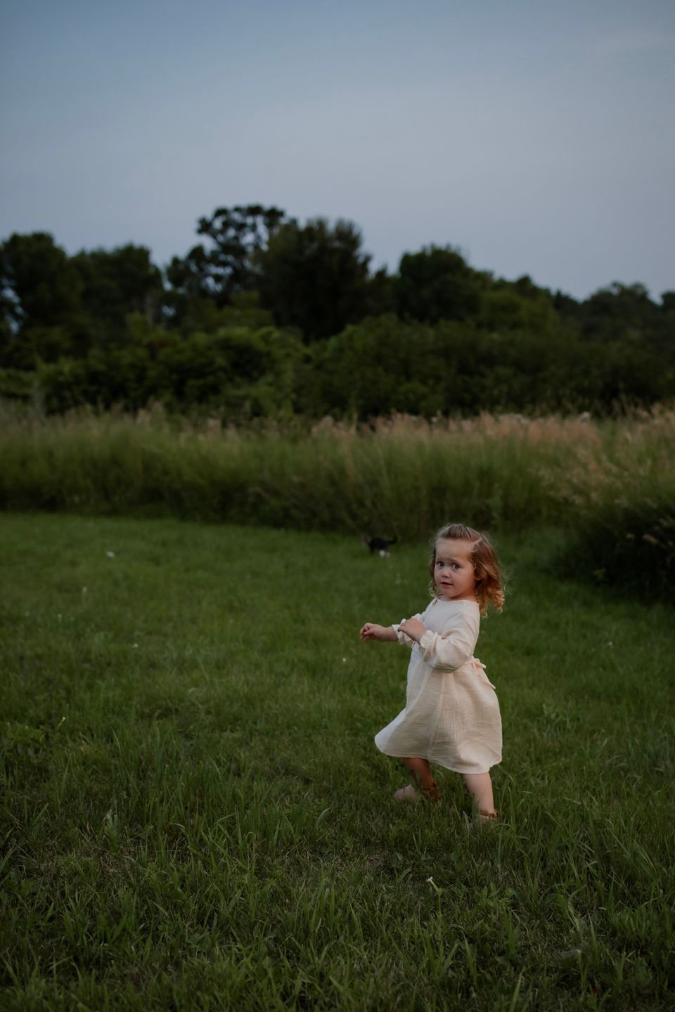 A person in a cream dress twirls in a grassy field at dusk.