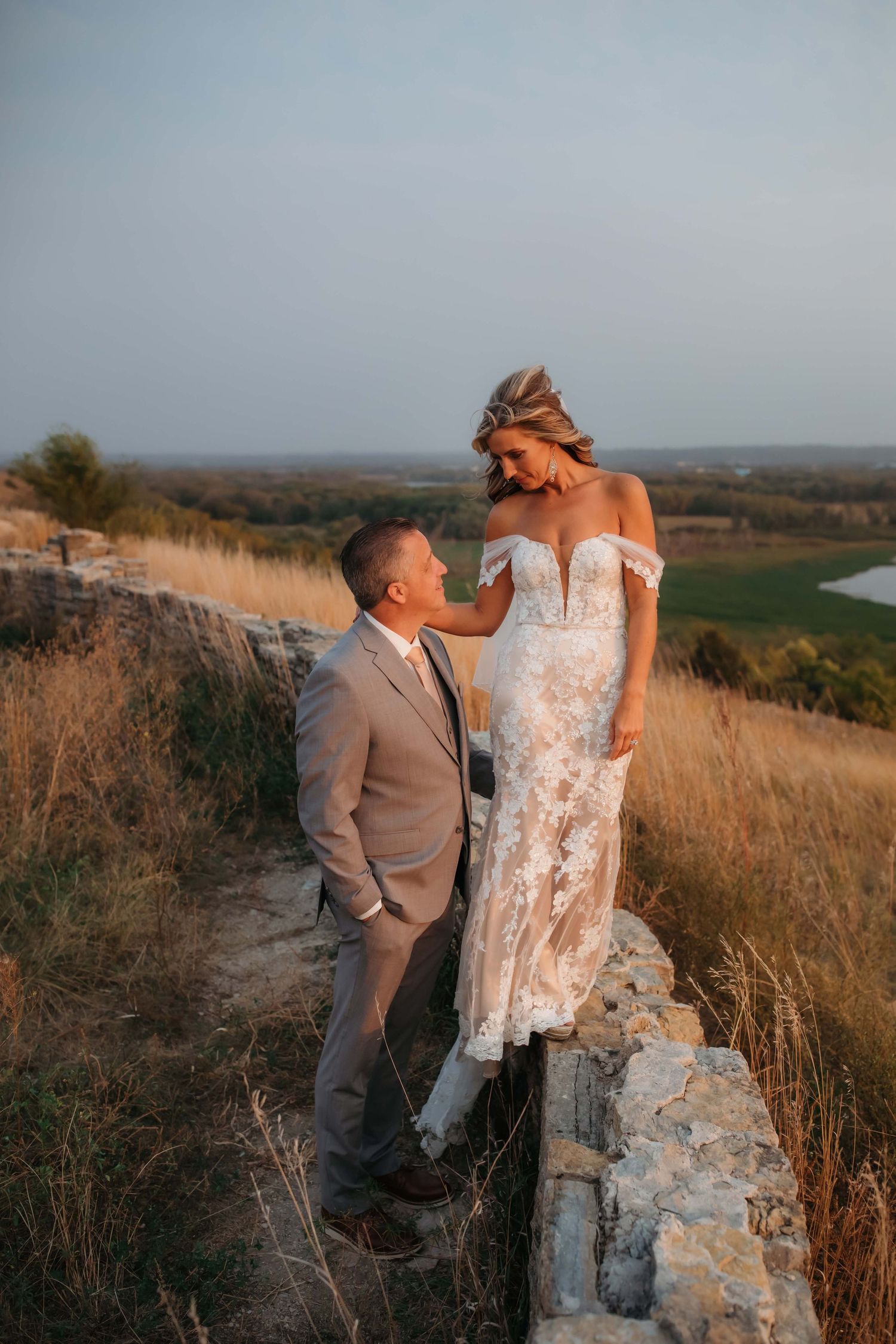 Couple shares intimate moment on stone wall overlooking scenic landscape at sunset.