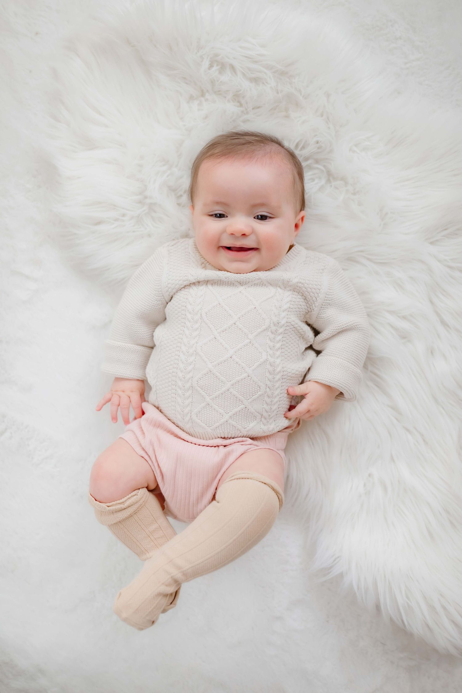 Baby in cream sweater and pink pants lying on white fur rug.