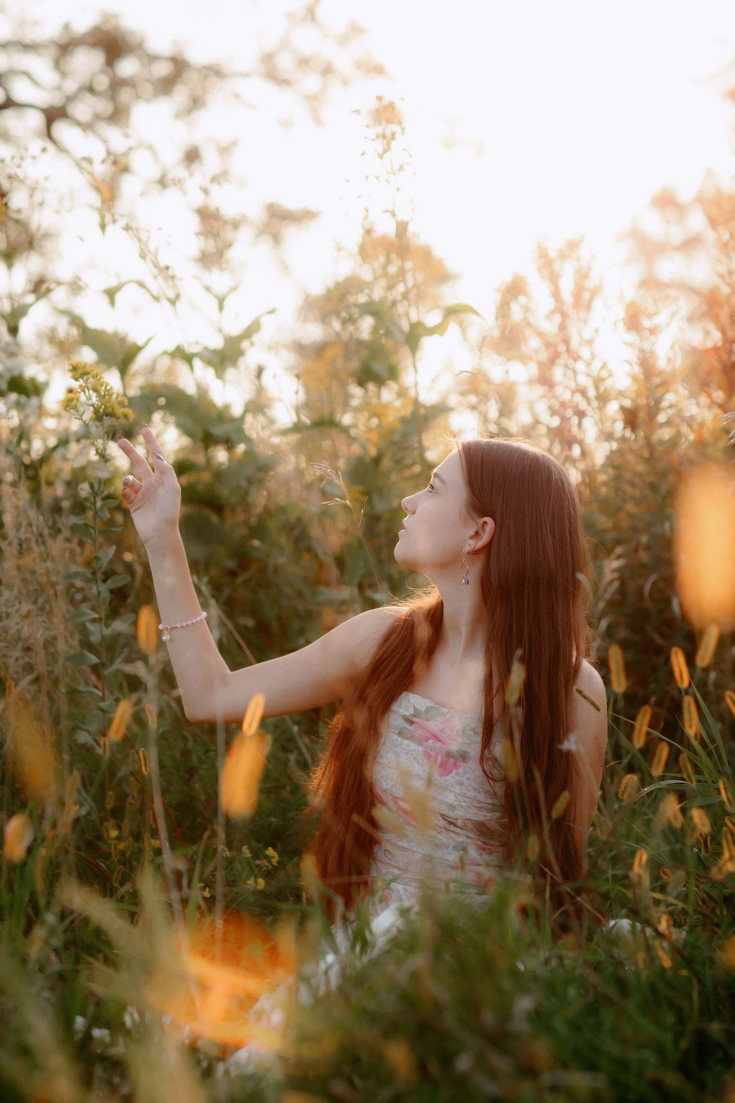 Long-haired figure in floral dress reaches toward sunlit sky in meadow of wildflowers.