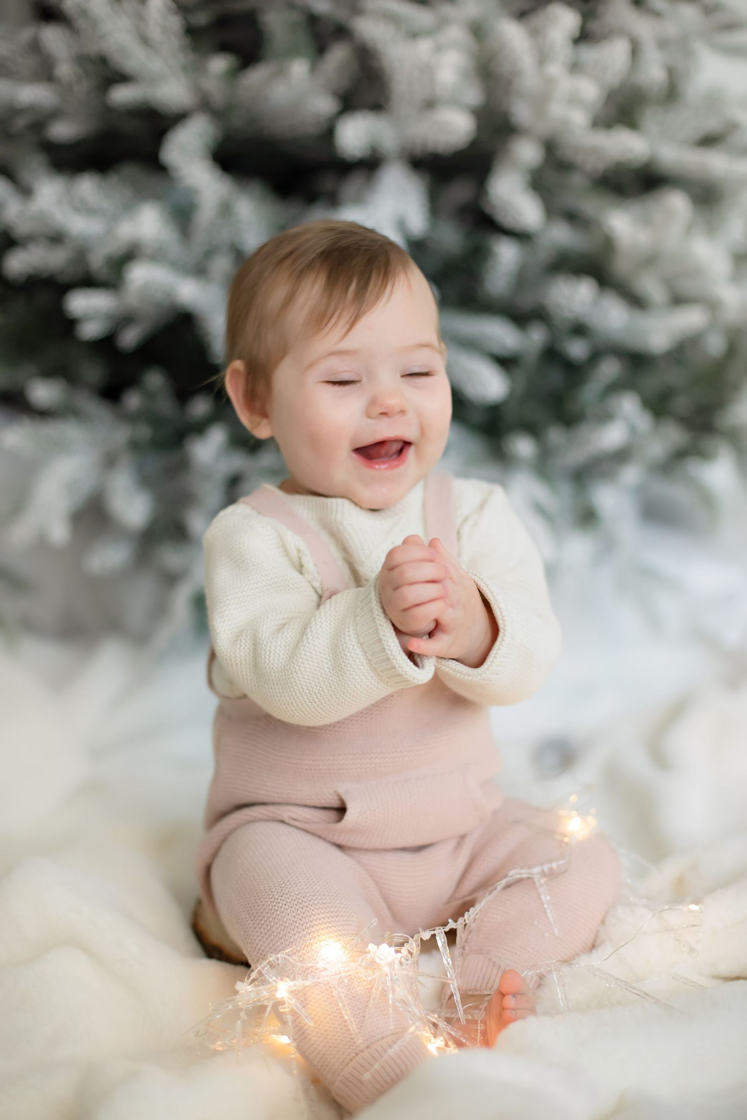 Baby in cream sweater and pink overalls sits happily with Christmas lights and snowy tree.