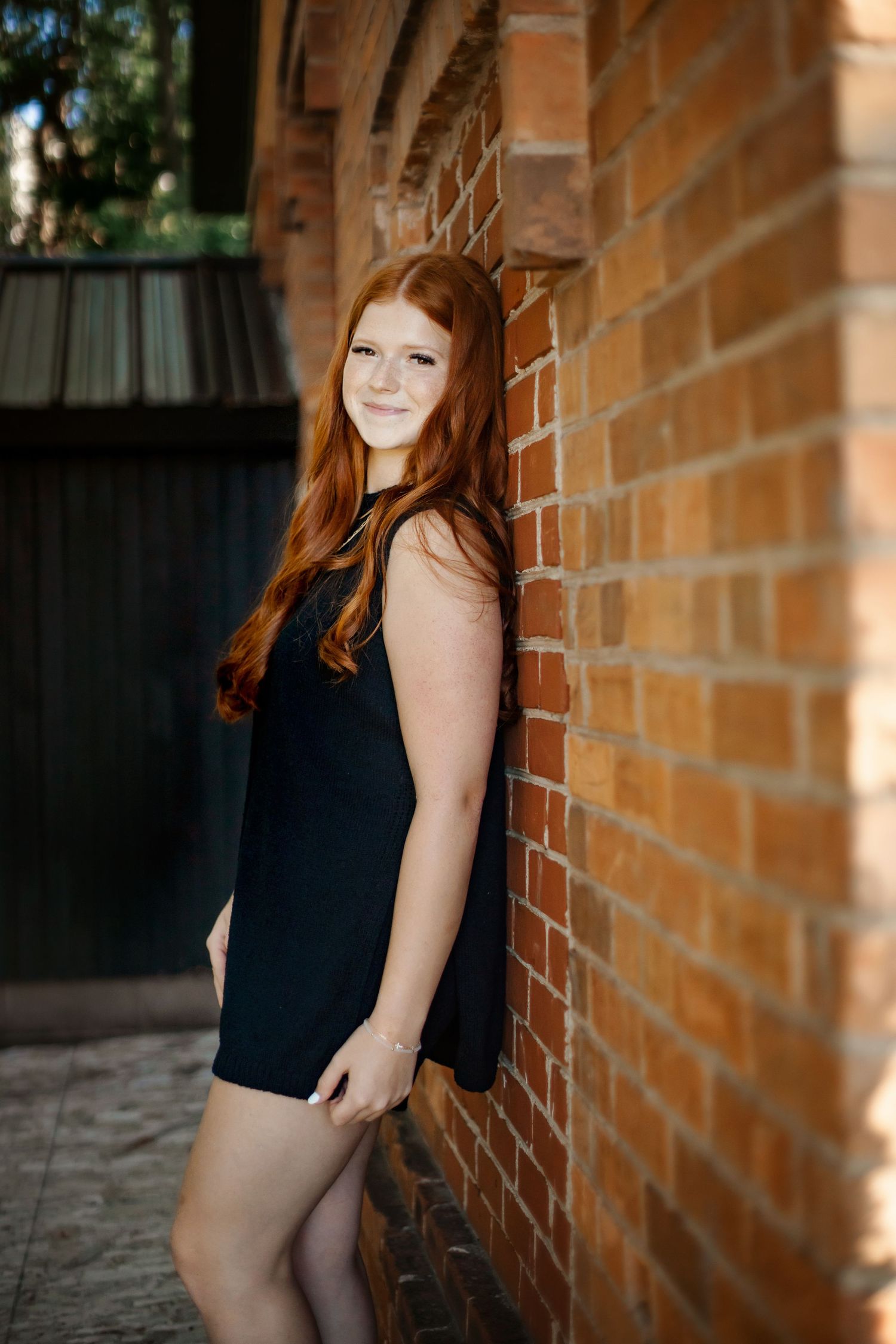 Portrait against brick wall featuring a black dress and long wavy red hair in natural lighting.