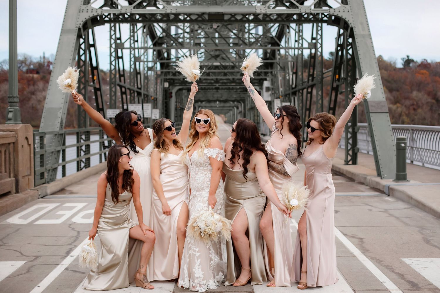Bridal party in champagne dresses celebrating joyfully on steel bridge.