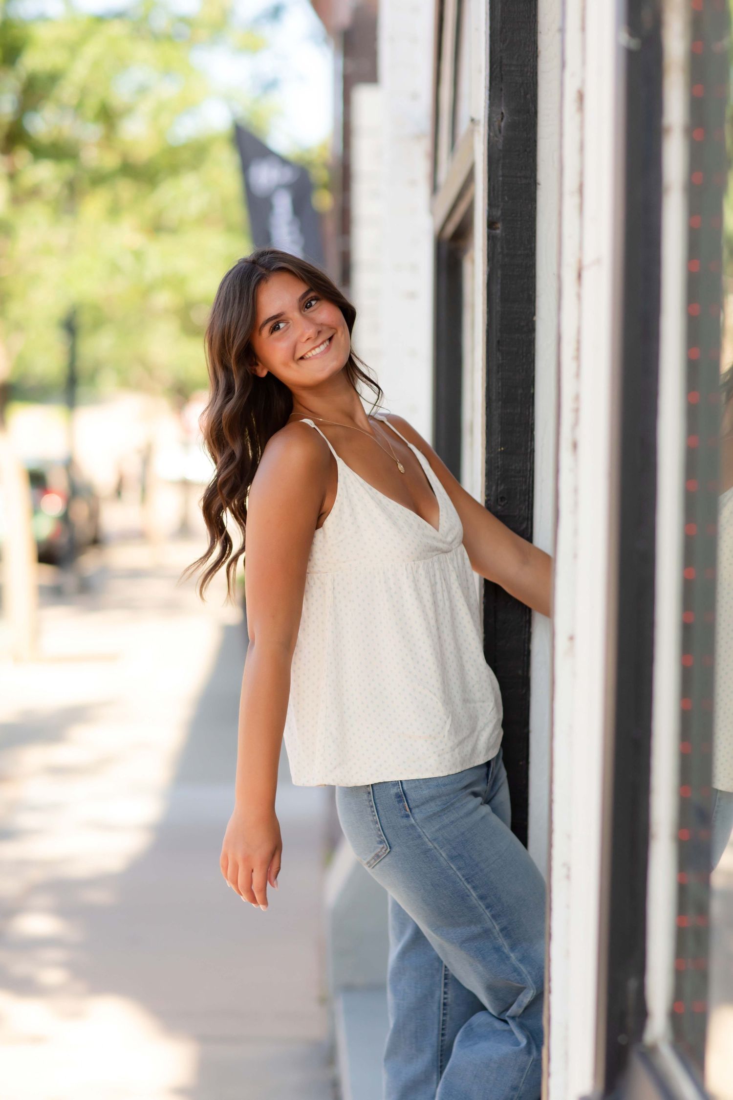 Young woman in white tank top and jeans leaning against building wall on sunny urban sidewalk, smiling.