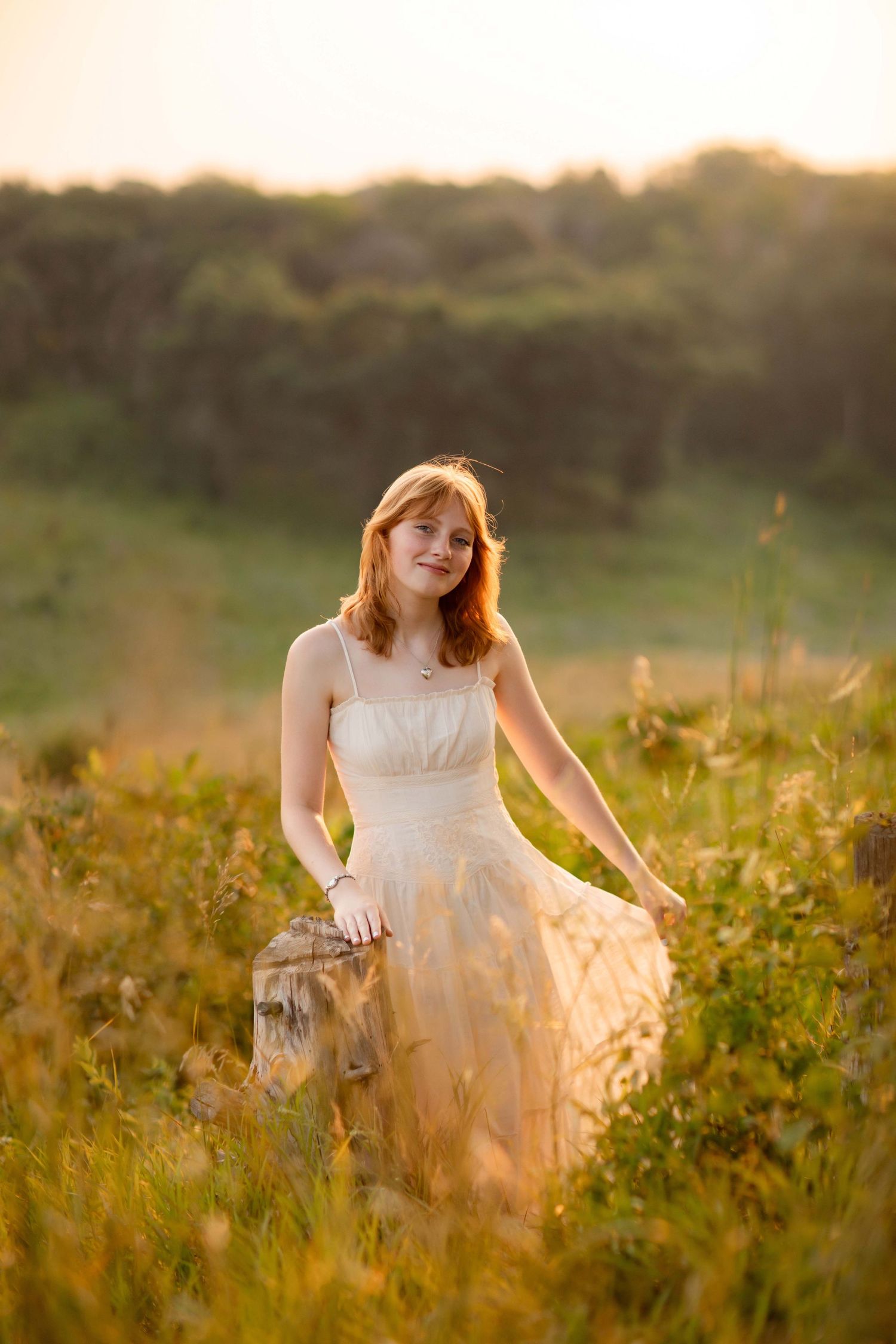 Young redhead woman in cream dress standing in golden sunlit meadow at sunset.