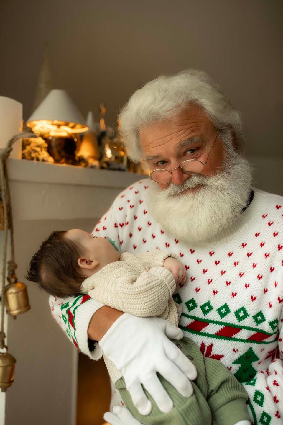 Santa Claus in festive sweater and white gloves gently holds a sleeping newborn baby near a decorated fireplace.