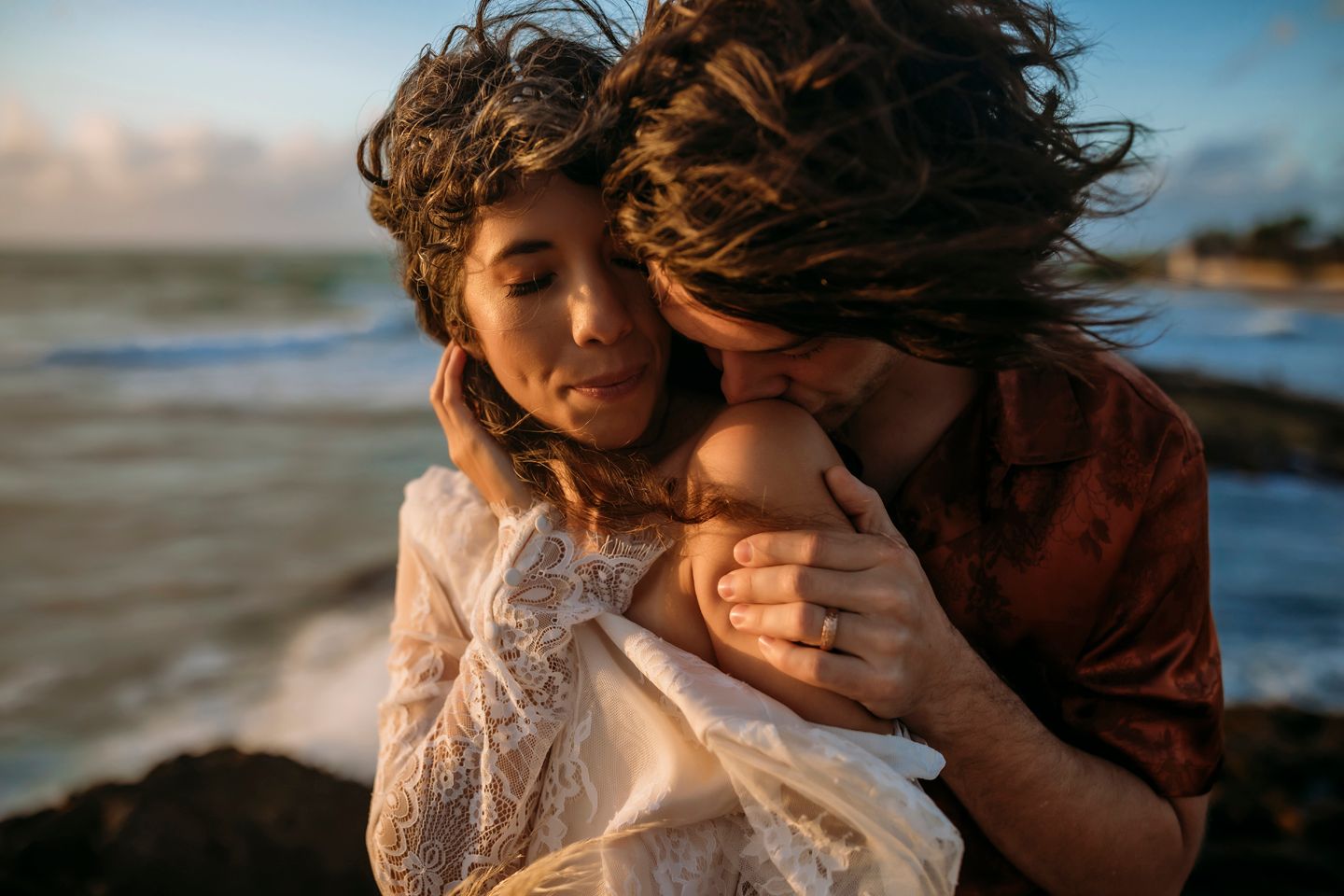 Romantic couple embraces on beach at sunset with windswept hair and lace dress details.