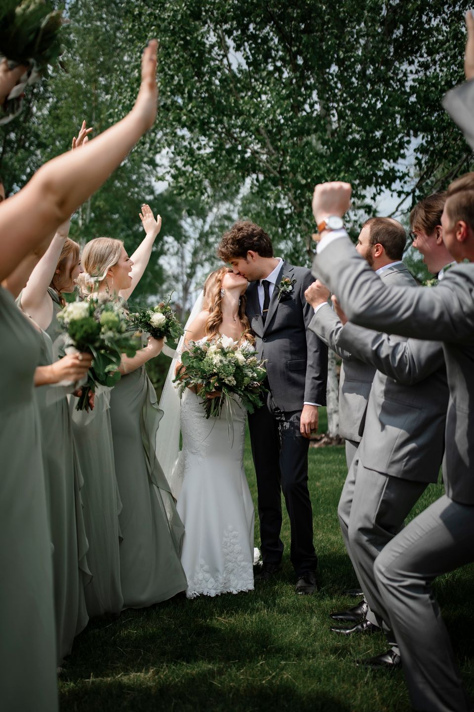 Wedding party in sage green and gray attire celebrates with raised hands during outdoor ceremony.