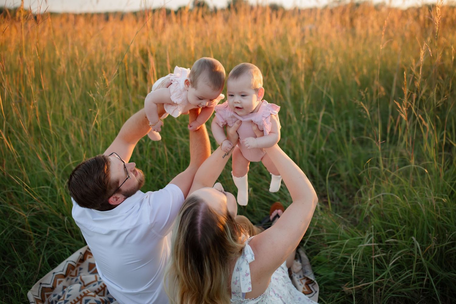 A family lifts twin babies overhead in a golden sunlit field at sunset.