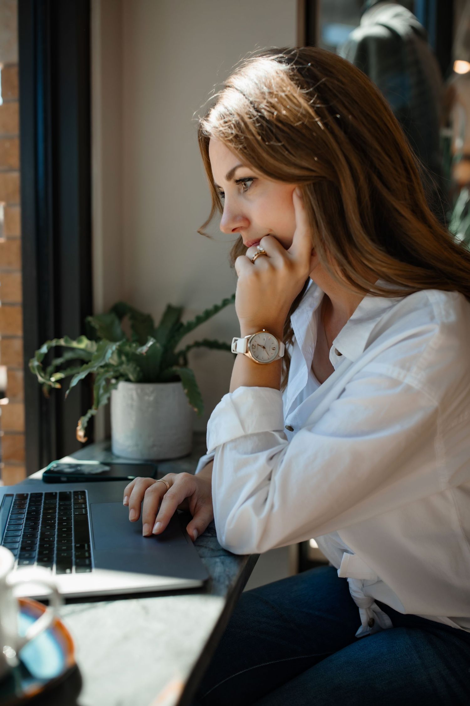 Side profile of someone working on laptop in white button-down shirt by window.