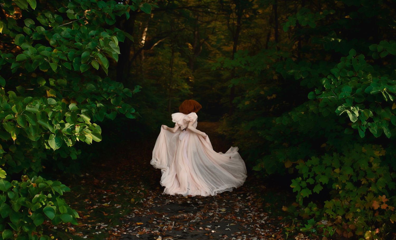 A flowing white dress swirls through a dark forest path lined with green foliage.