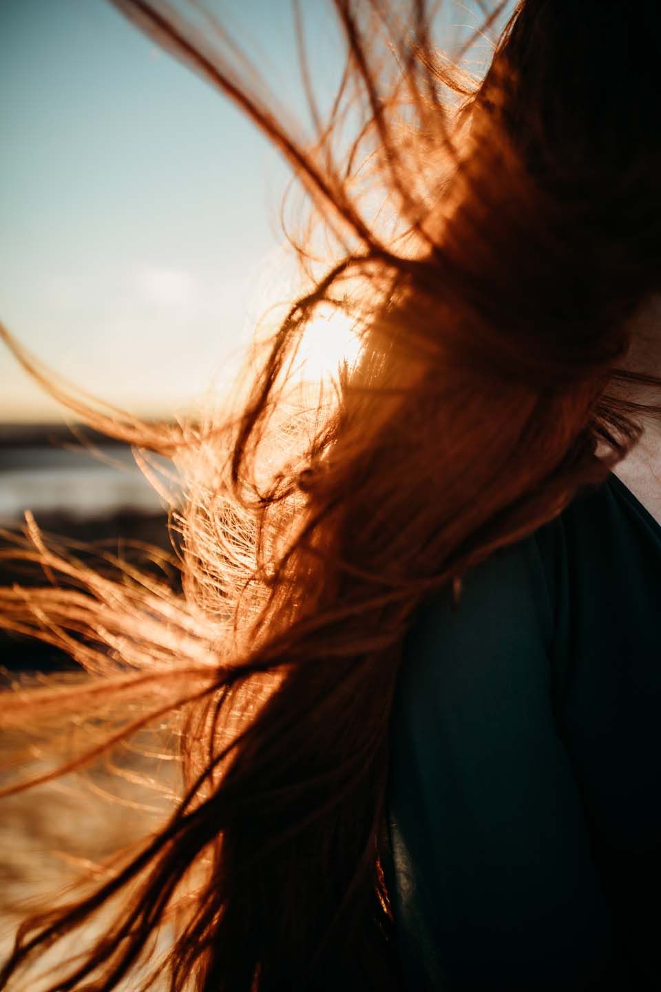 Long hair flowing dramatically in the wind against a sunset sky and ocean backdrop.