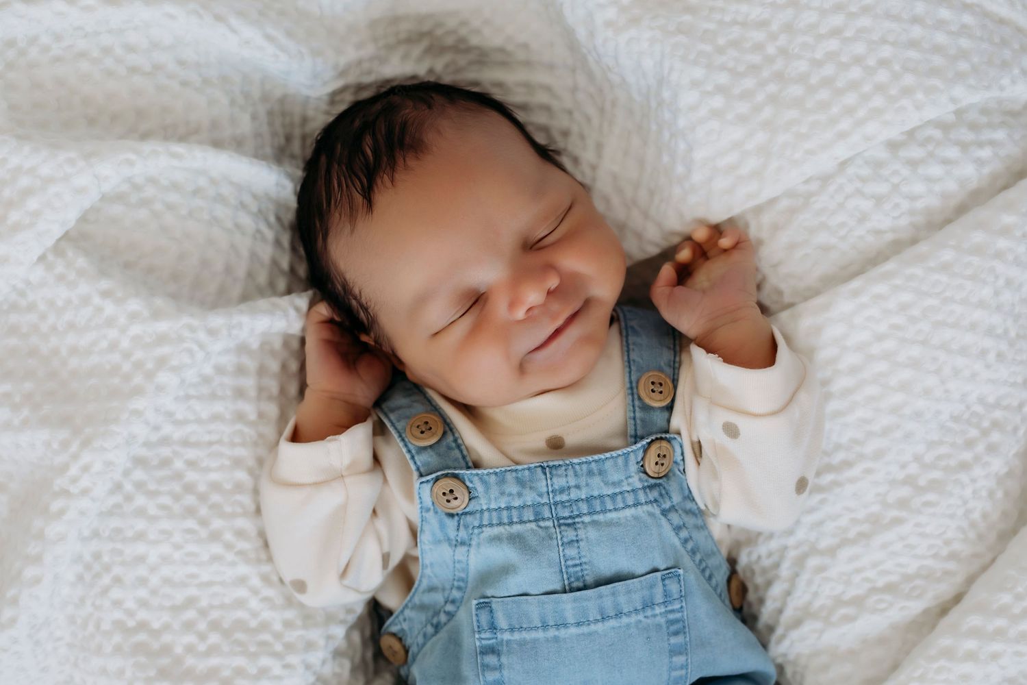 Sweet newborn smiling while sleeping in blue overalls on white textured blanket.