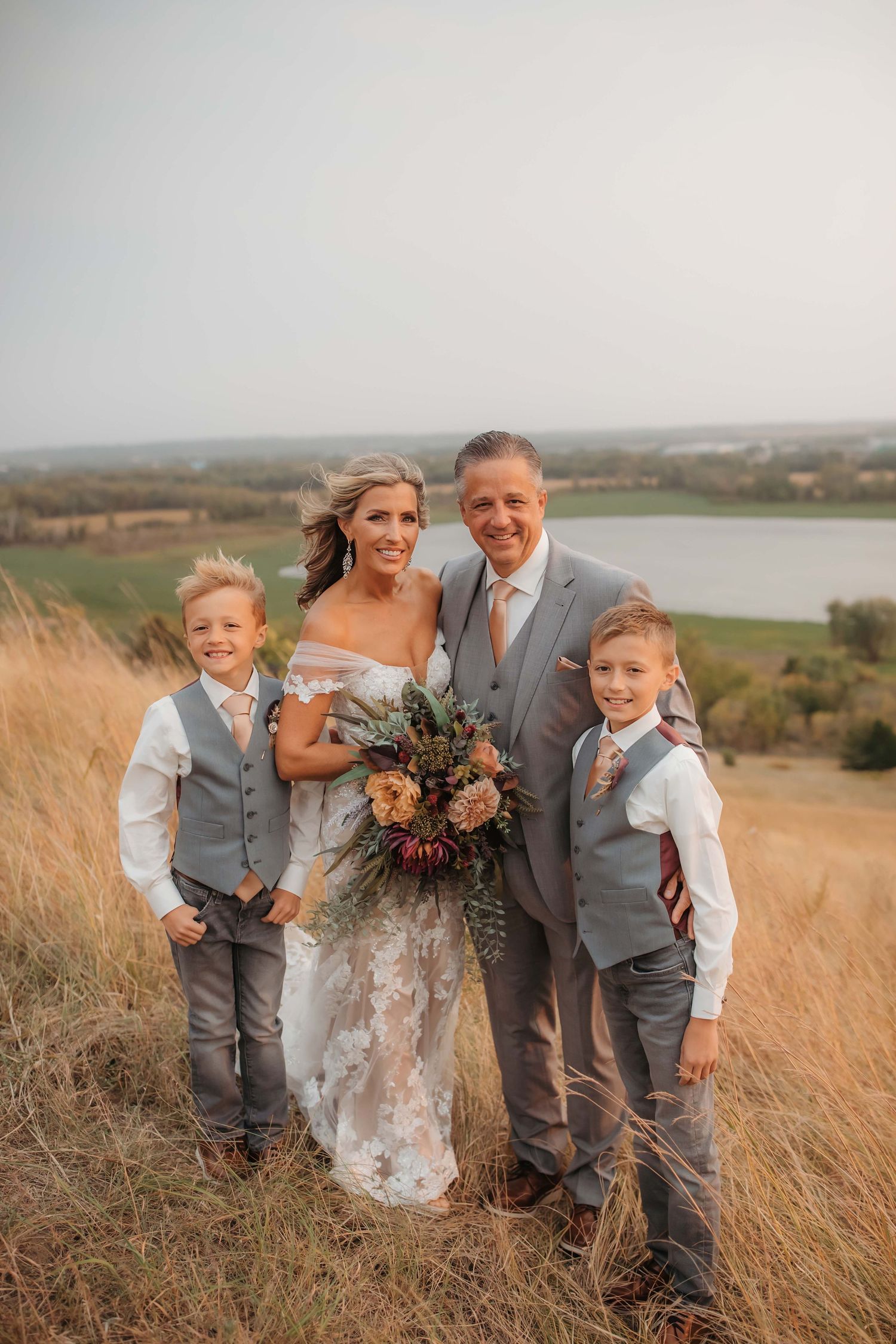 Wedding portrait of family standing in tall grass with lake view at sunset.
