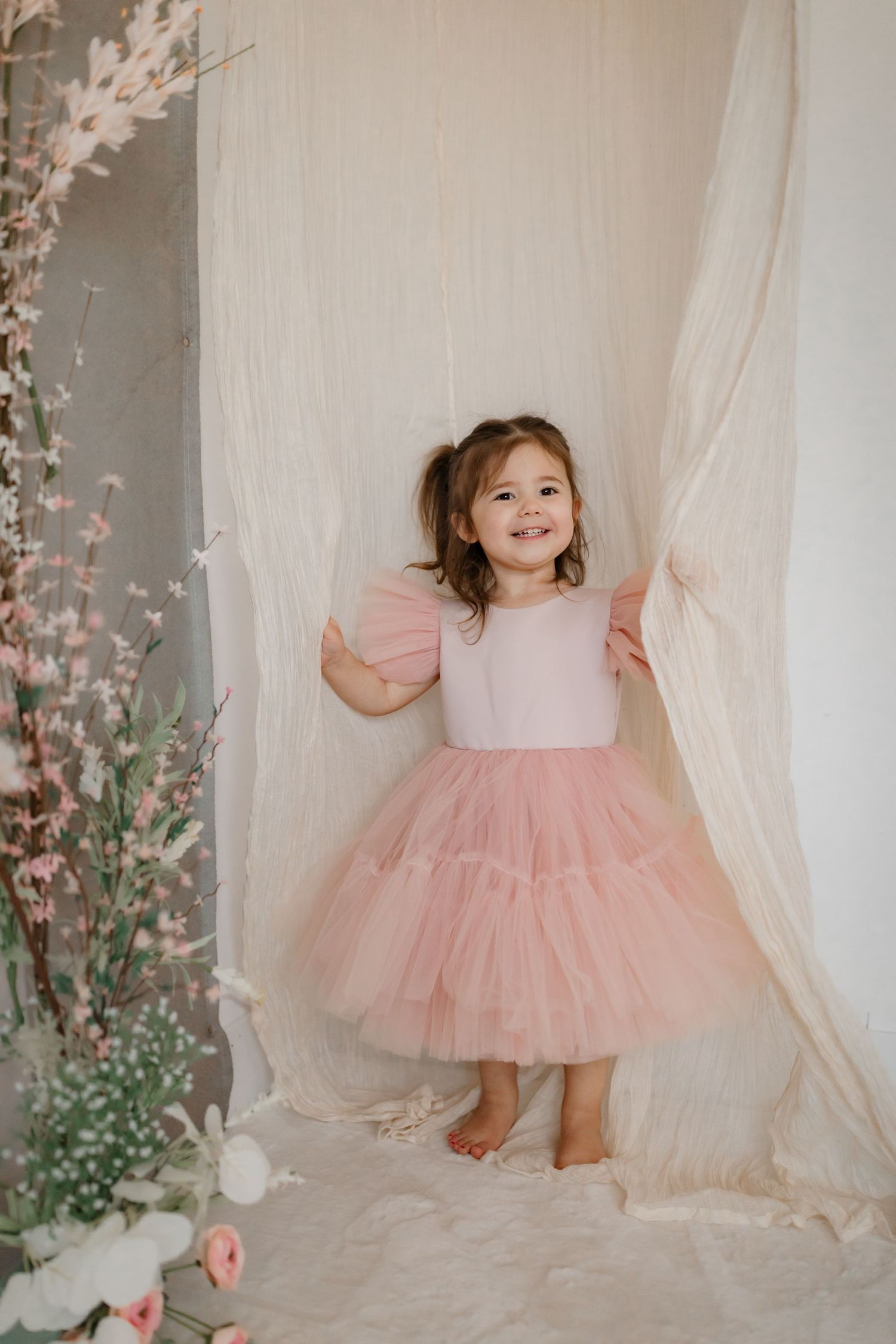 Child in pink tulle dress stands between flowing white curtains and cherry blossoms.