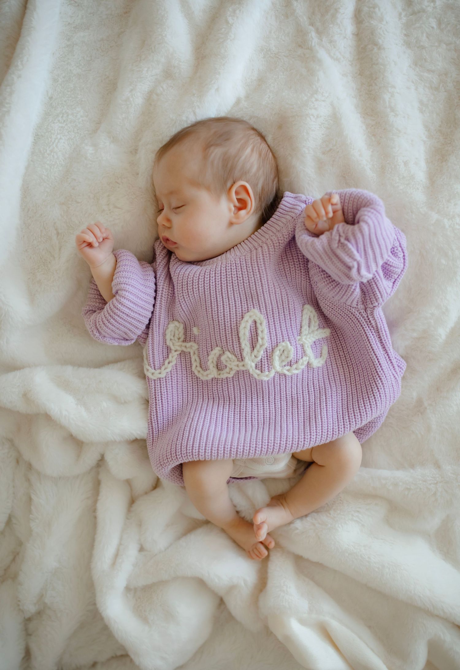 Peaceful baby in lavender knit sweater with embroidered text resting on soft white fabric.