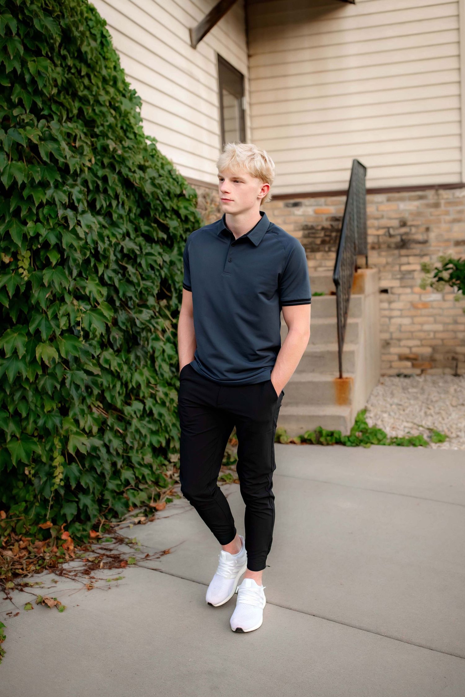 Standing pose by ivy-covered wall wearing dark polo shirt and white shoes.