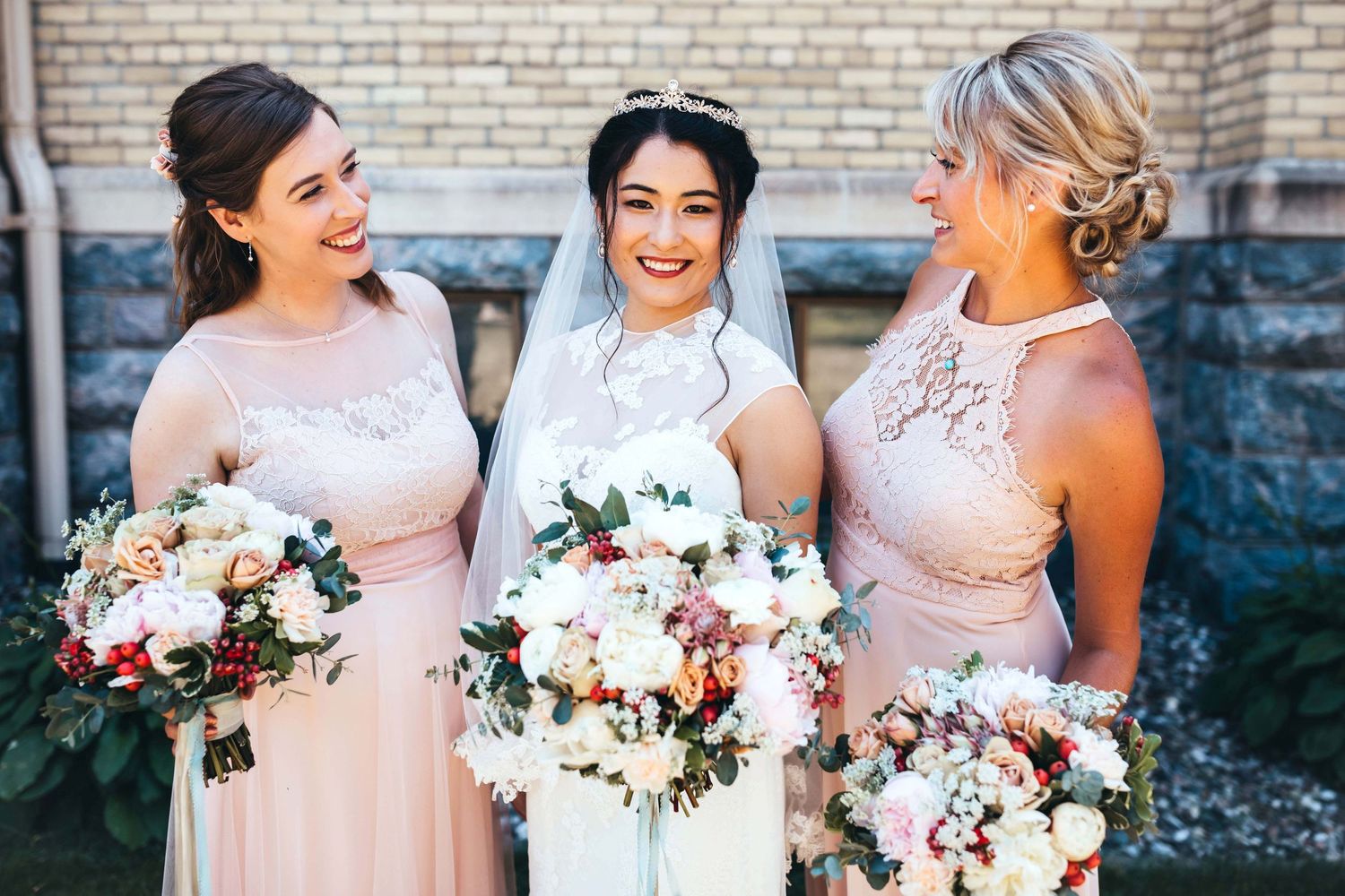 Three bridesmaids in blush pink dresses hold elegant blush and white bouquets while smiling together.