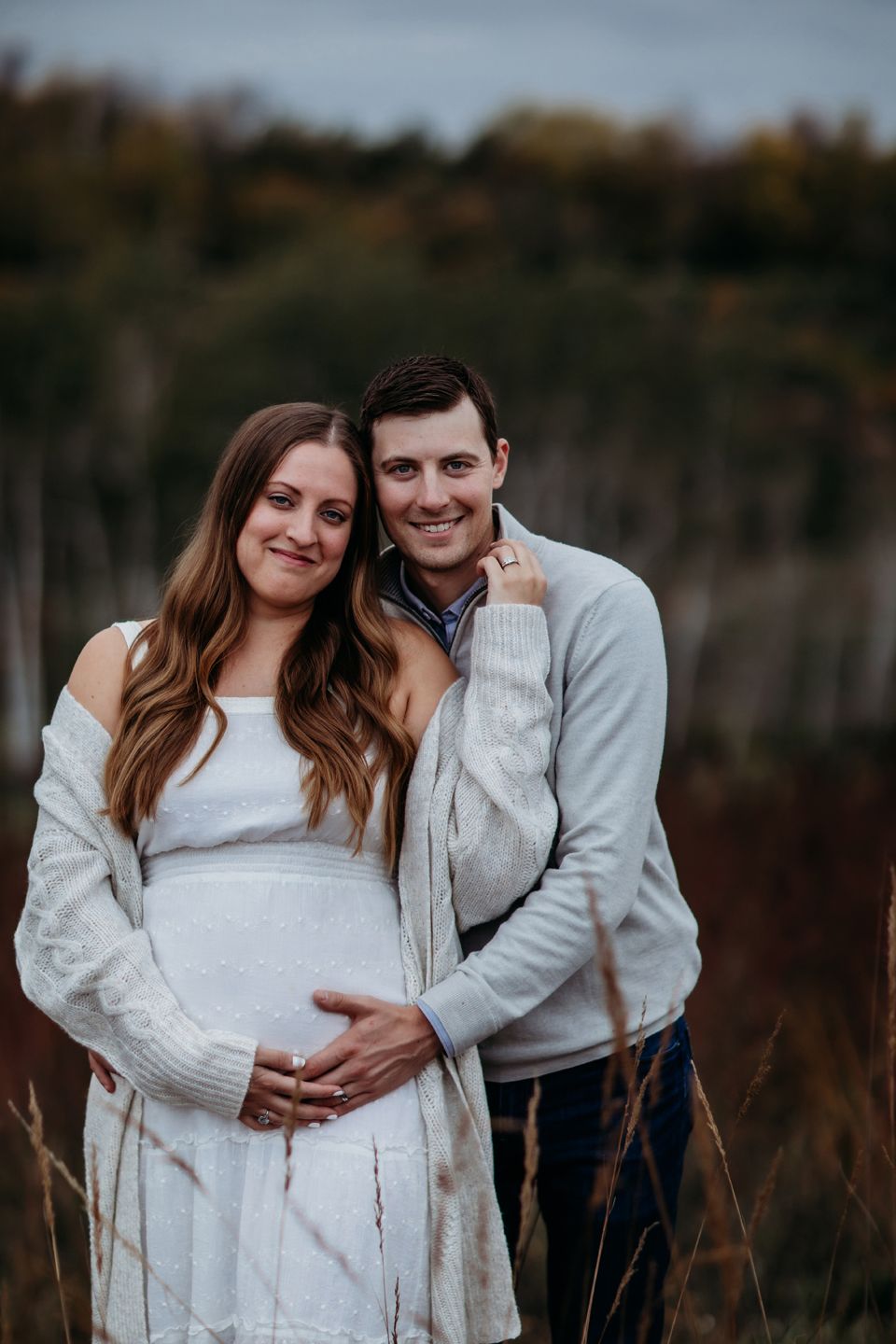 A couple embraces during a maternity photoshoot outdoors in autumn, wearing cozy light-colored sweaters.