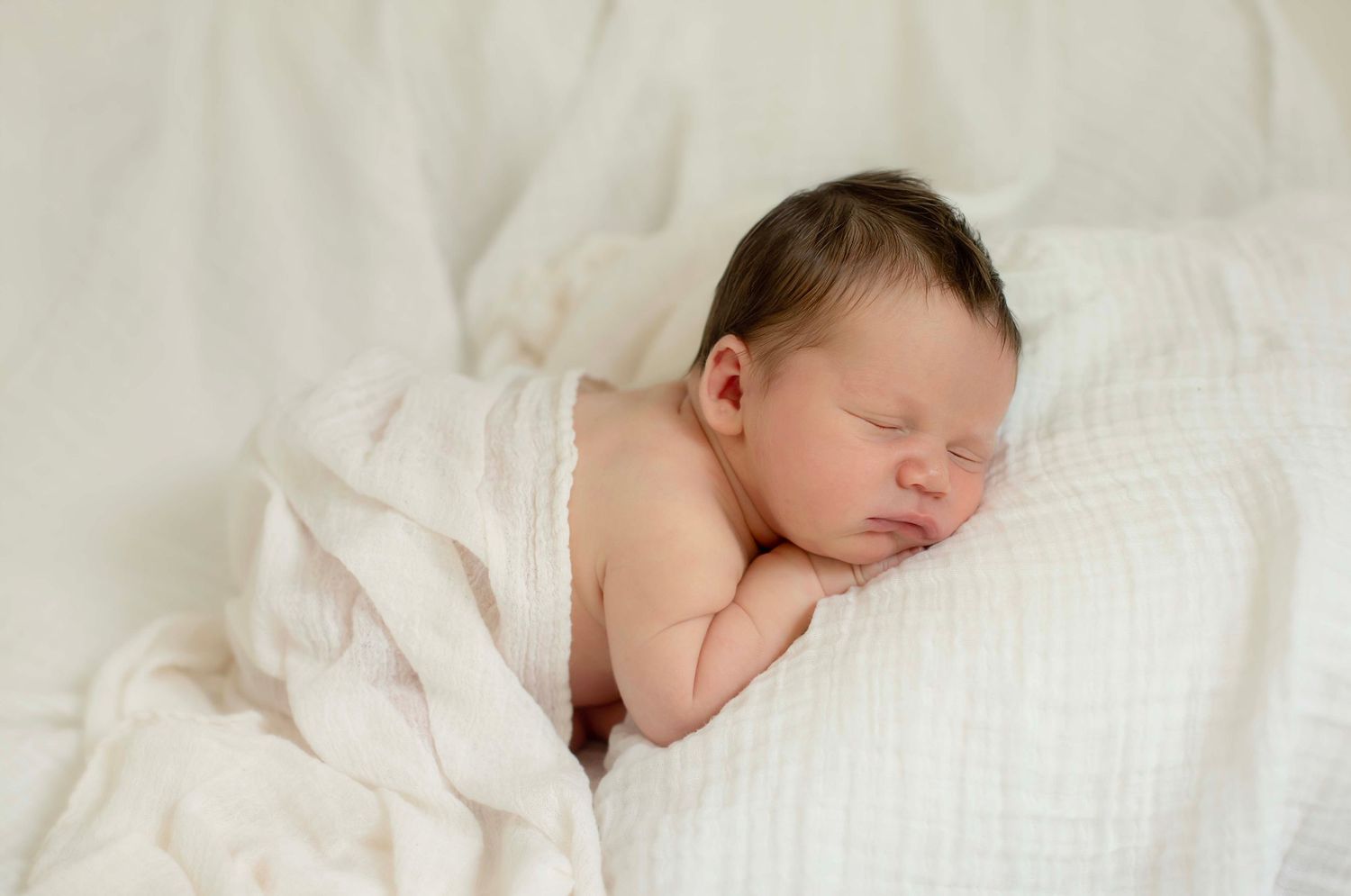 Peaceful newborn baby sleeping on white pillow wrapped in soft white muslin blanket in professional photo.
