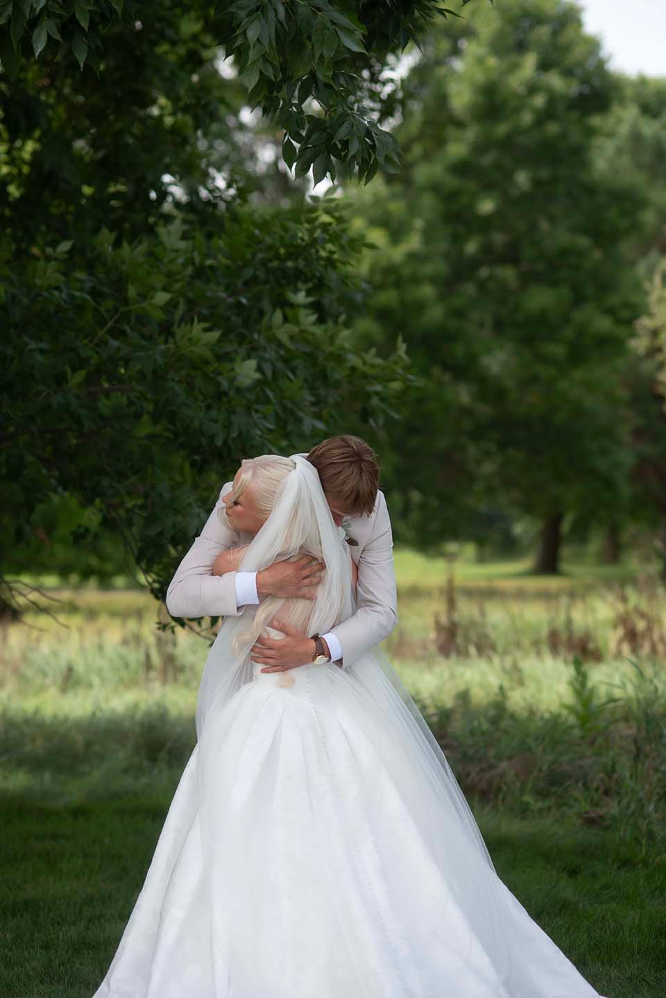 A bride and groom share a romantic embrace outdoors, surrounded by lush green trees on their wedding day.