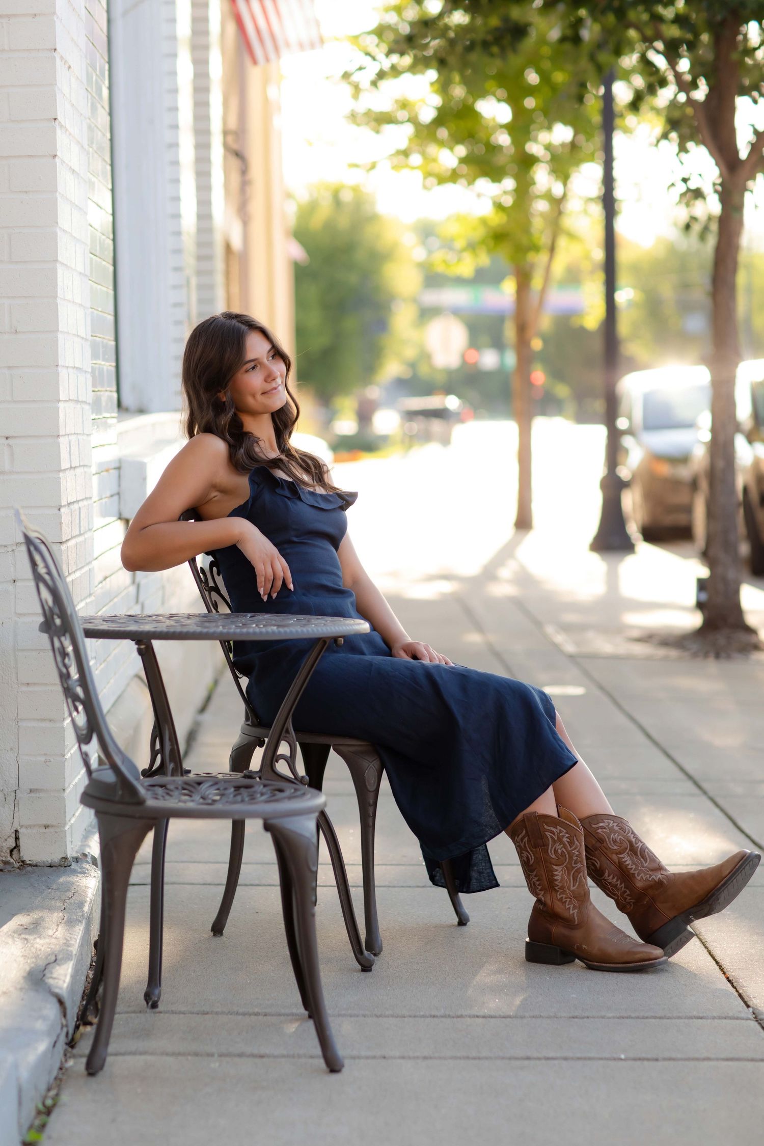 Young woman in navy jumpsuit and cowboy boots sits relaxed at outdoor cafe table on sunny urban sidewalk.