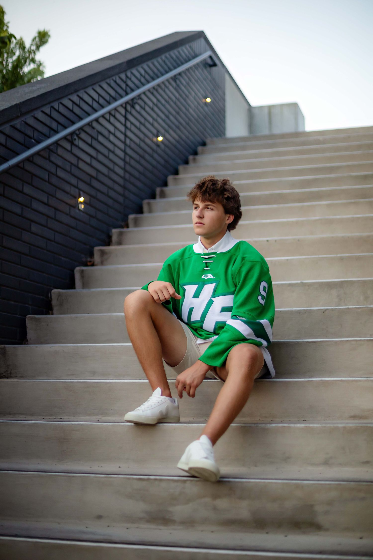 Teen boy in green hockey jersey sitting on outdoor concrete steps at dusk in white sneakers.