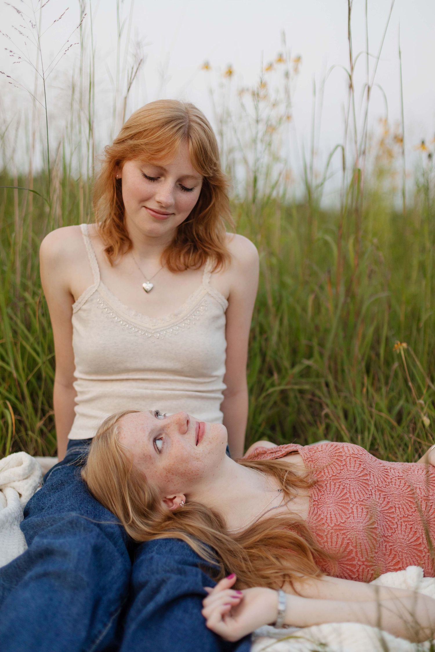 Two young redhead women relaxing outdoors in tall grass, one lying down, sharing a peaceful moment together.