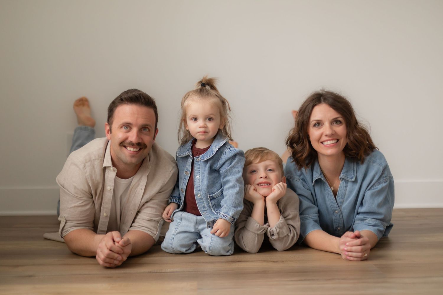 Family members lying on floor in casual clothing for portrait session in neutral toned studio setting.