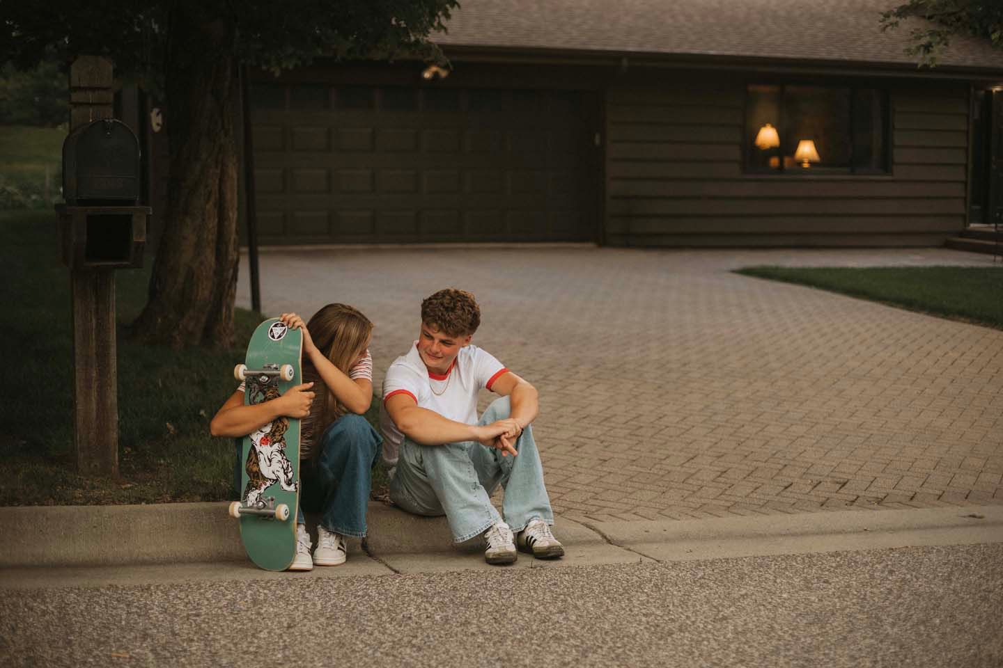 Two teens sit on a curb sharing a moment, one holding a green skateboard, in front of a house at dusk.