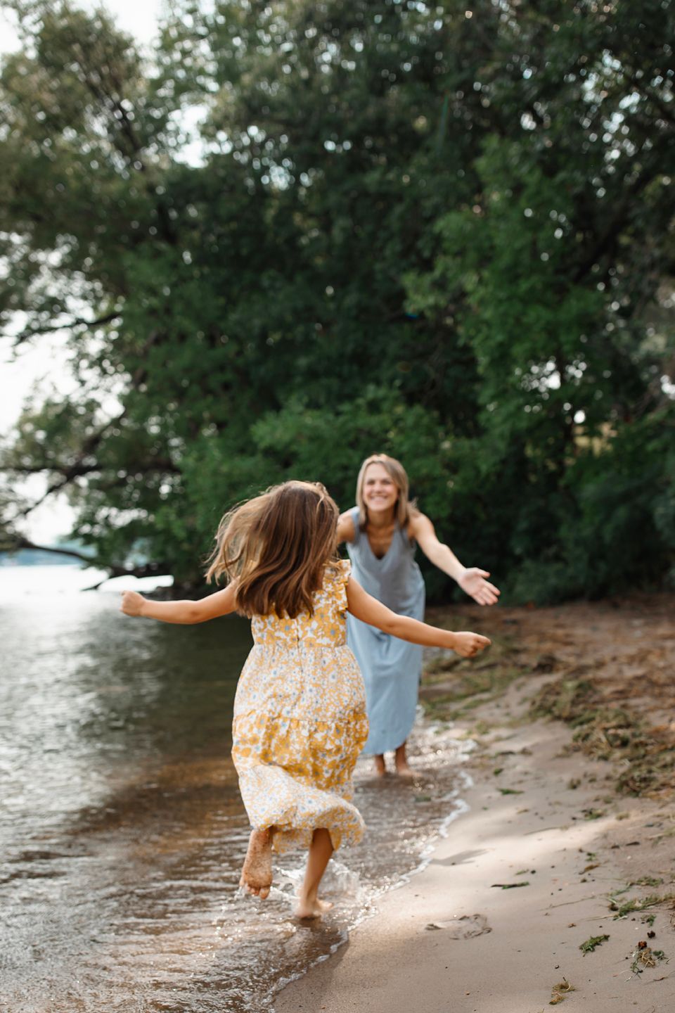 Two people in flowy dresses run and dance joyfully along a sandy beach shoreline by a tree-lined lake.