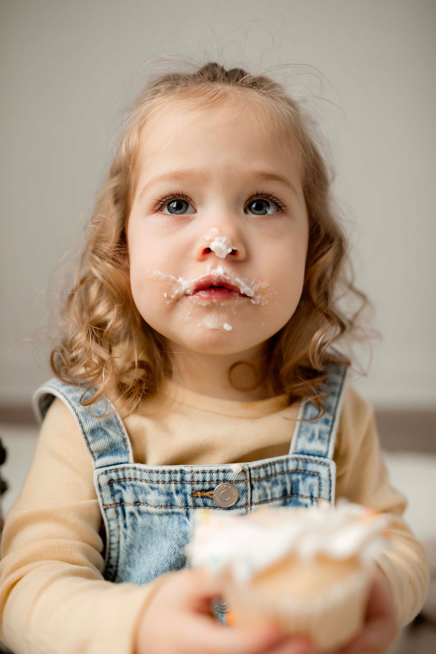 Toddler in denim overalls has whipped cream on their nose and mouth while enjoying a sweet treat.