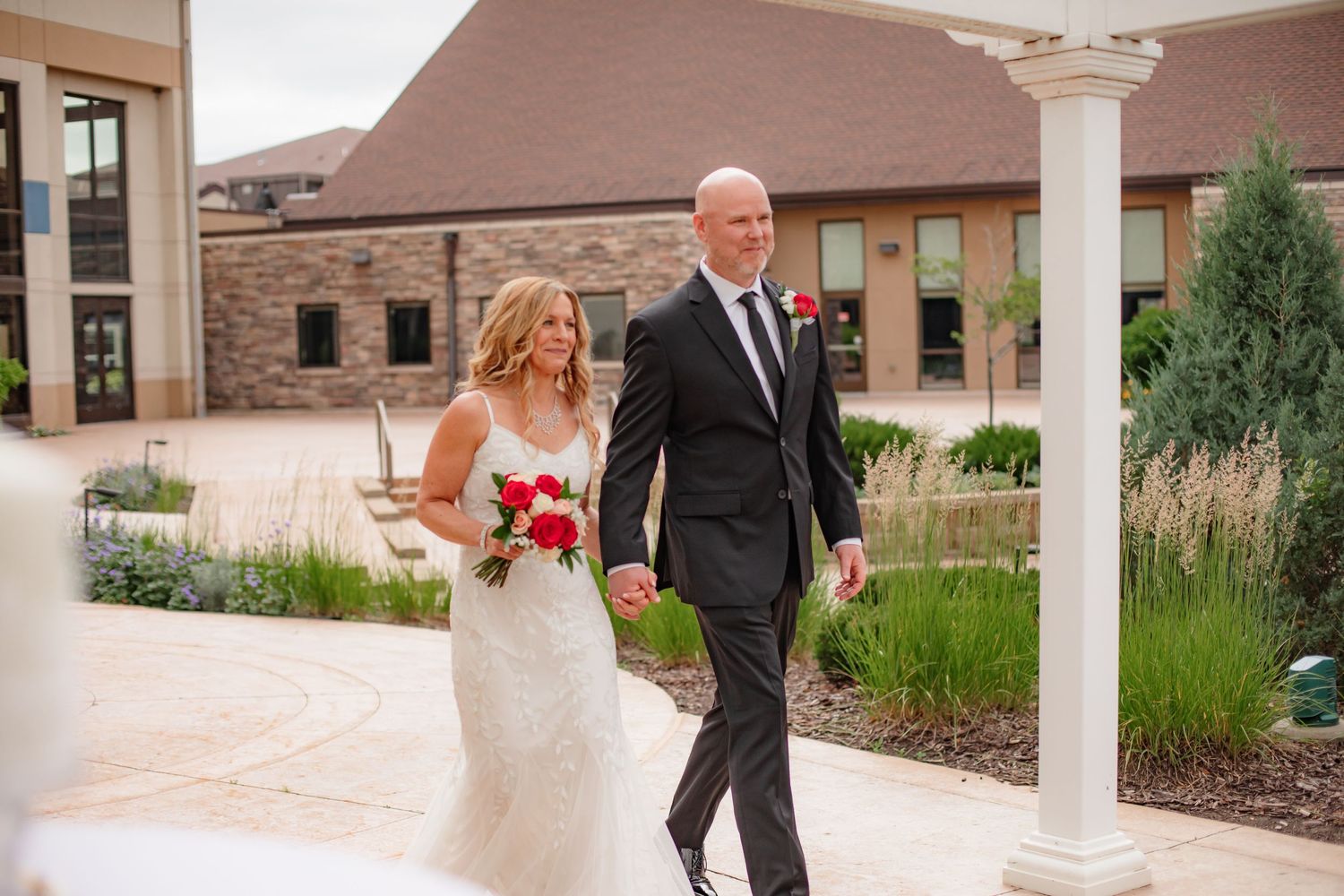 A bride in a fitted lace gown and groom in a black suit walk together outside a stone building with white columns.