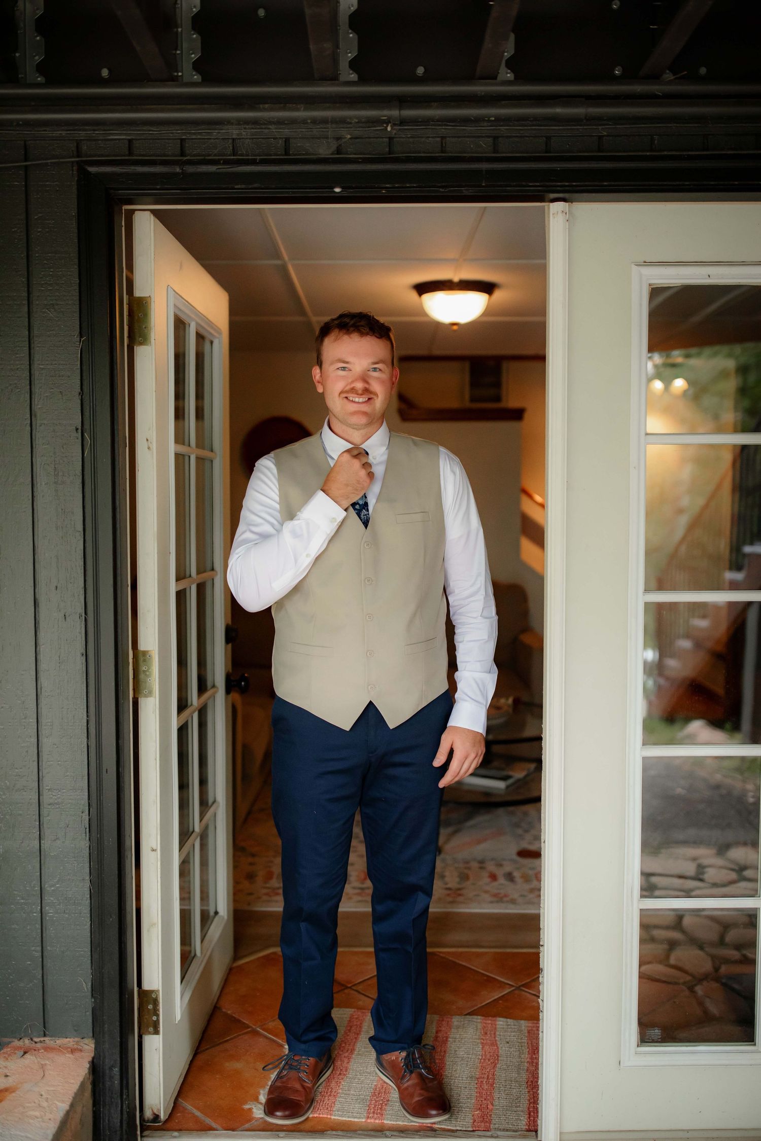 A groom in a light beige vest and navy pants poses for photos in a doorway sequence.